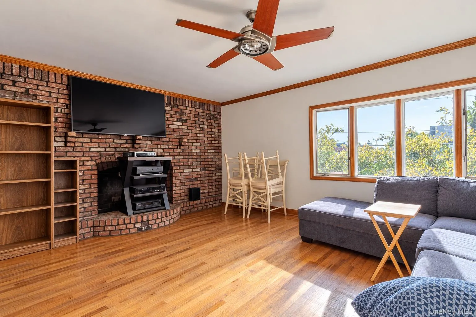 Exposed brick fireplace, wood-finish flooring, ceiling fan, built-in shelving, and large windows with wood-trim casing Exposed brick fireplace, wood-finish flooring, ceiling fan, built-in shelving, and large windows with wood-trim casing