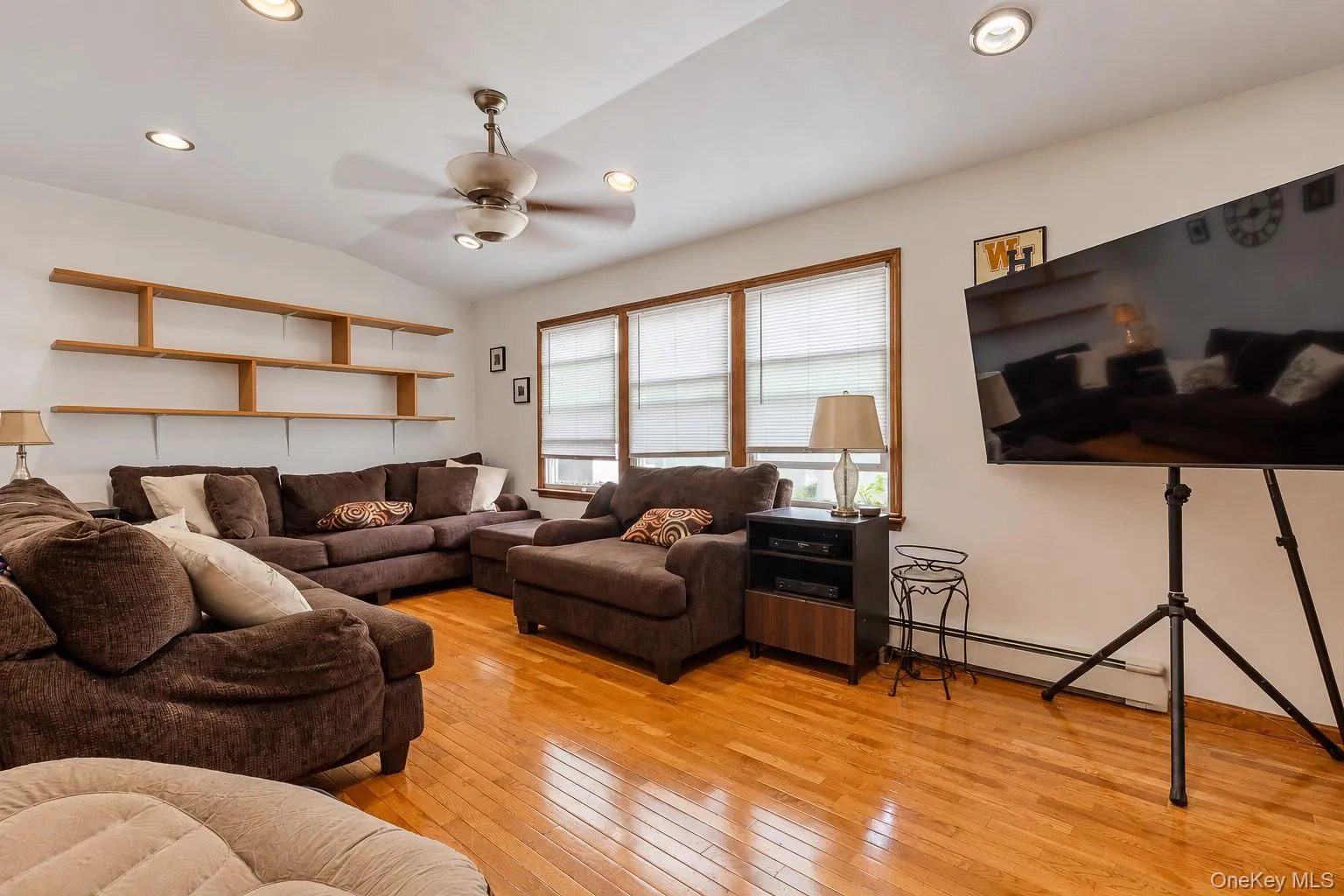 Living area featuring wood-finish flooring, recessed lighting, a ceiling fan, tiered built-in shelving, and multiple windows with wood-trimmed casings Living area featuring wood-finish flooring, recessed lighting, a ceiling fan, tiered built-in shelving, and multiple windows with wood-trimmed casings