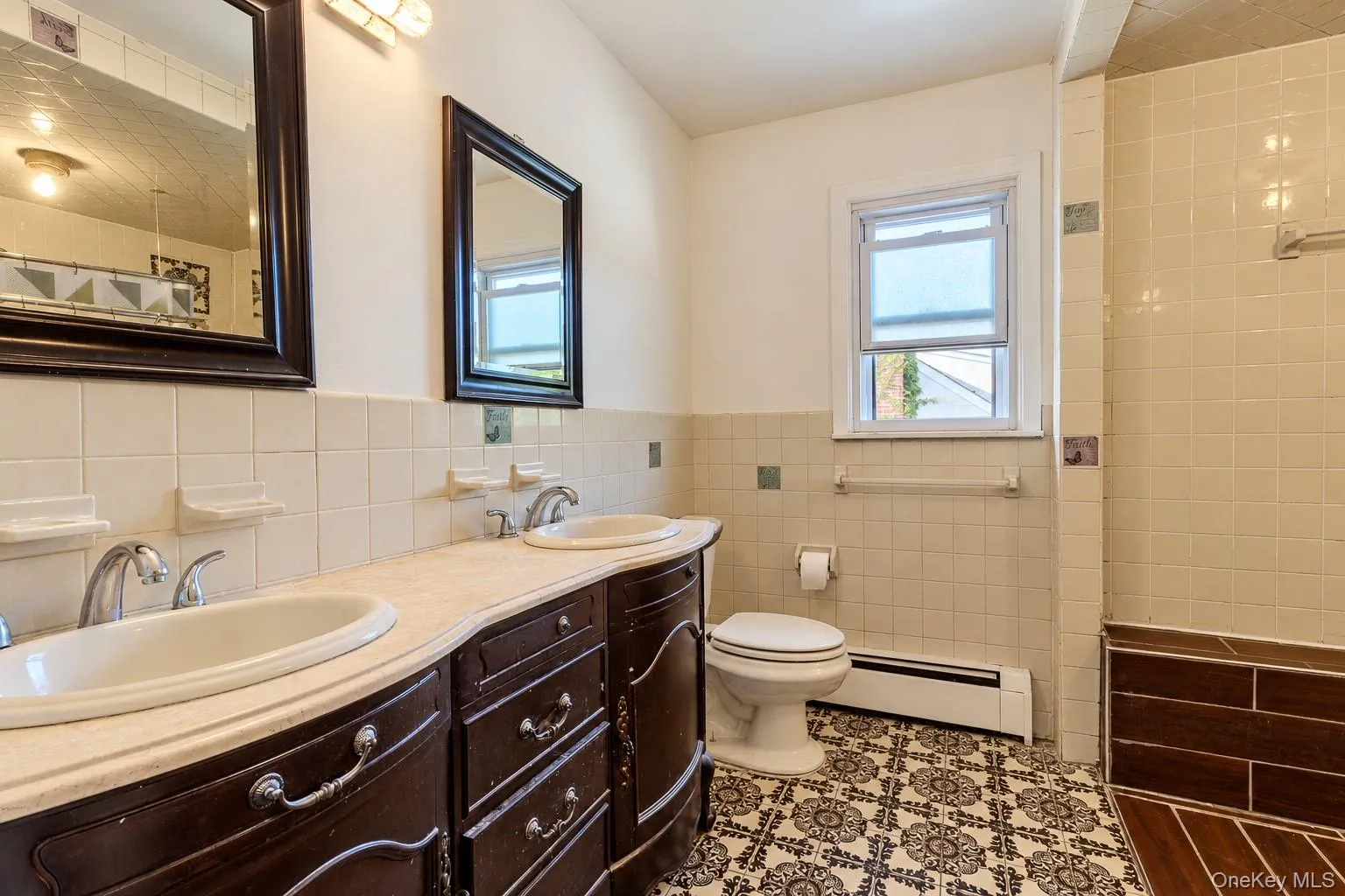 Bathroom featuring a dual-sink vanity with light-toned countertop, ornate dark-finish cabinetry, decorative tile flooring, and a tiled shower surround with a built-in bench Bathroom featuring a dual-sink vanity with light-toned countertop, ornate dark-finish cabinetry, decorative tile flooring, and a tiled shower surround with a built-in bench