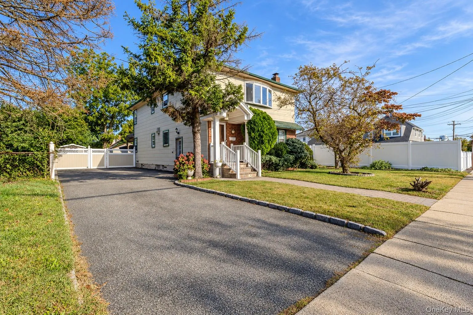 Two-story residence featuring light-toned siding, a covered entryway with white railings, an asphalt driveway, and a white privacy fence Two-story residence featuring light-toned siding, a covered entryway with white railings, an asphalt driveway, and a white privacy fence