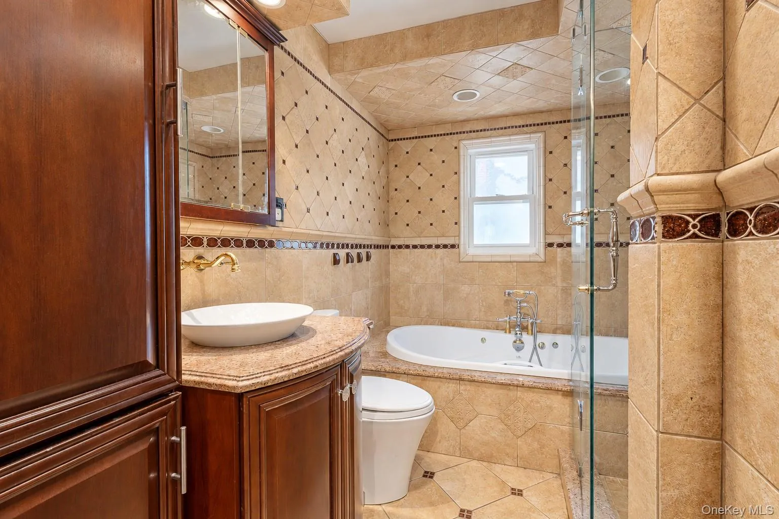 Bathroom featuring stone tile walls with diamond-pattern detailing, a built-in jetted tub, glass-enclosed shower, wood-finish vanity with a vessel sink, and metallic wall-mounted fixtures Bathroom featuring stone tile walls with diamond-pattern detailing, a built-in jetted tub, glass-enclosed shower, wood-finish vanity with a vessel sink, and metallic wall-mounted fixtures