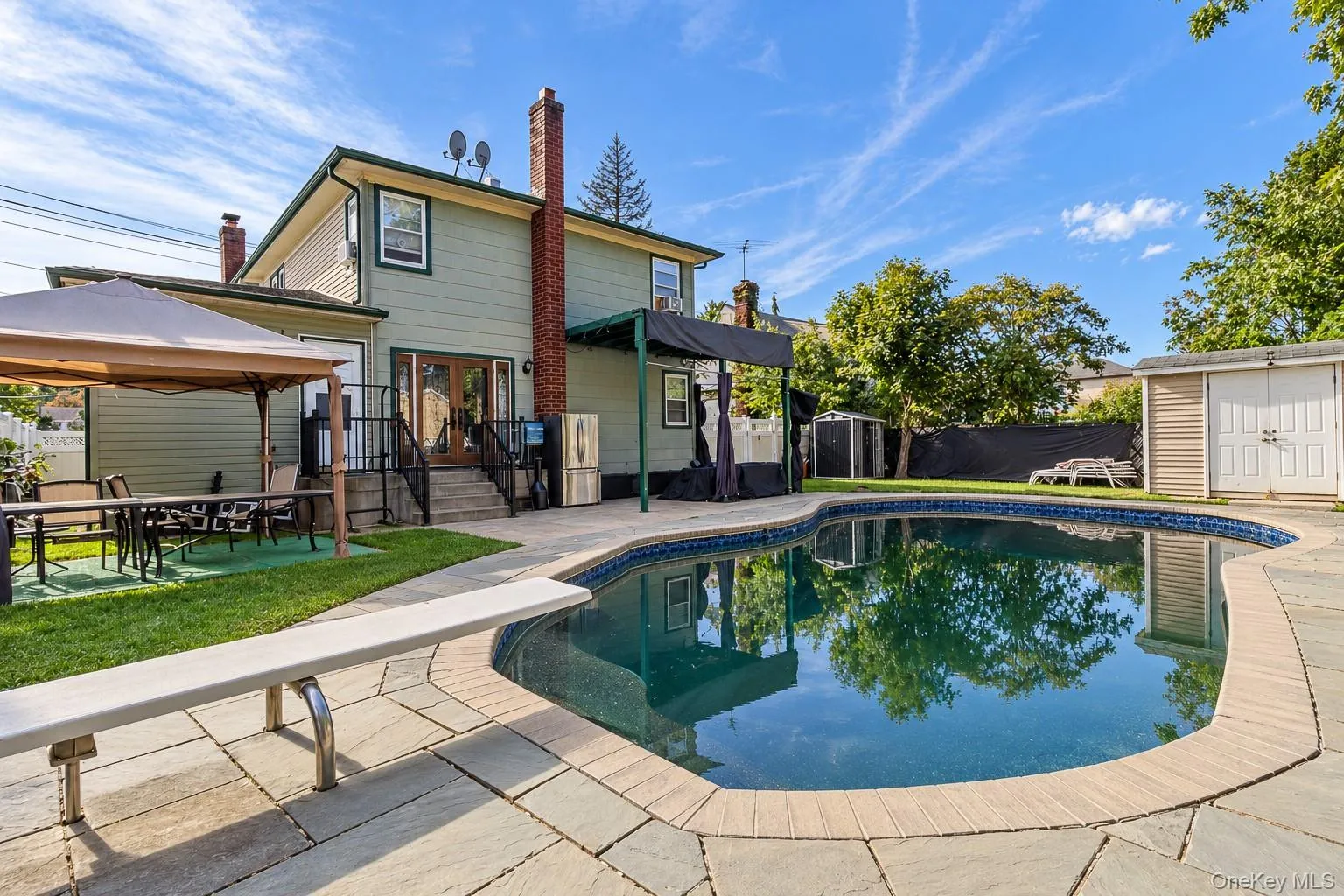 Free-form in-ground swimming pool with a diving board, surrounded by stone patio hardscaping, external brick chimney, and a detached storage structure Free-form in-ground swimming pool with a diving board, surrounded by stone patio hardscaping, external brick chimney, and a detached storage structure