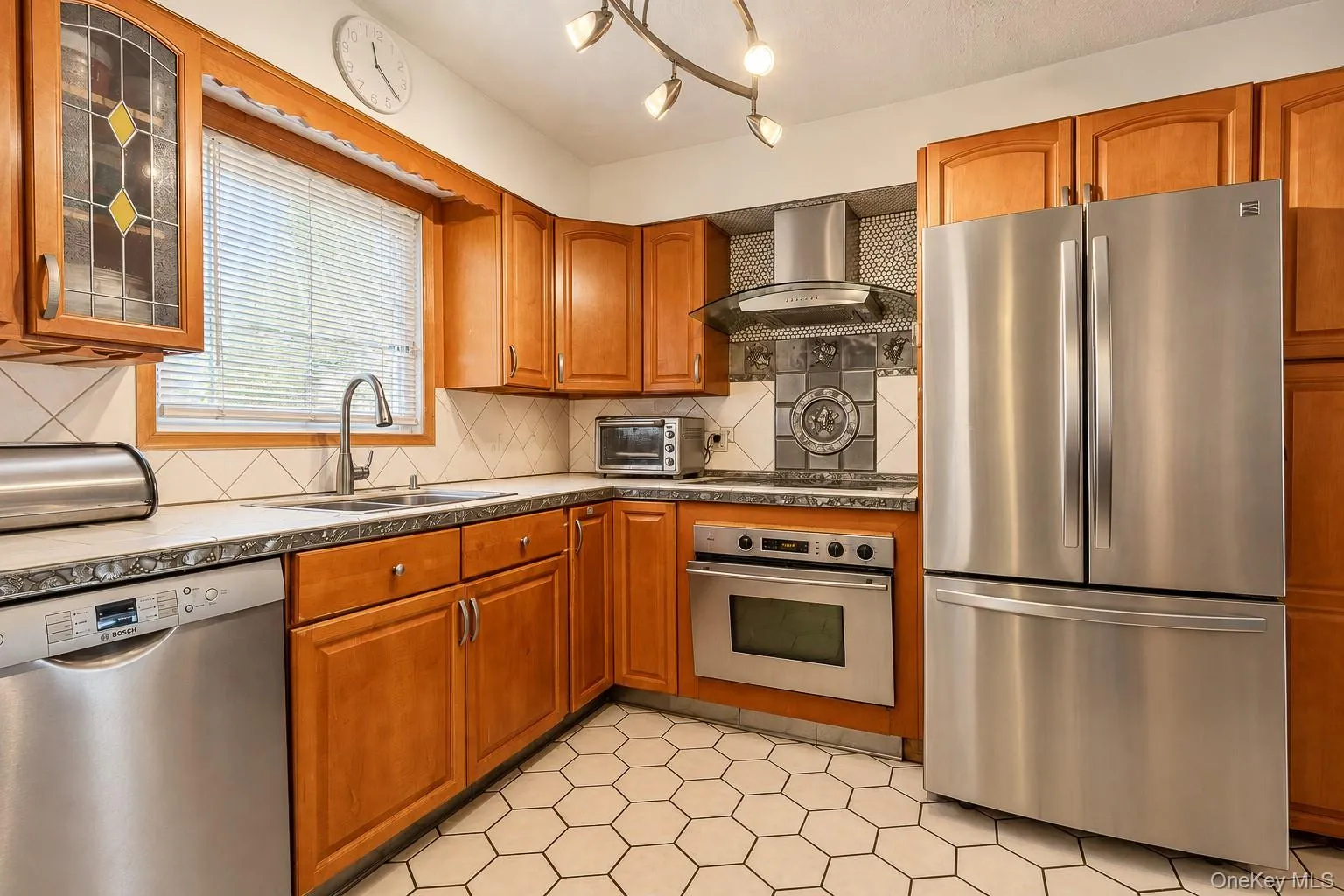 Kitchen featuring custom wood cabinetry, stainless steel appliances, a modern chimney-style range hood, hexagonal tile flooring, and a decorative tile backsplash Kitchen featuring custom wood cabinetry, stainless steel appliances, a modern chimney-style range hood, hexagonal tile flooring, and a decorative tile backsplash