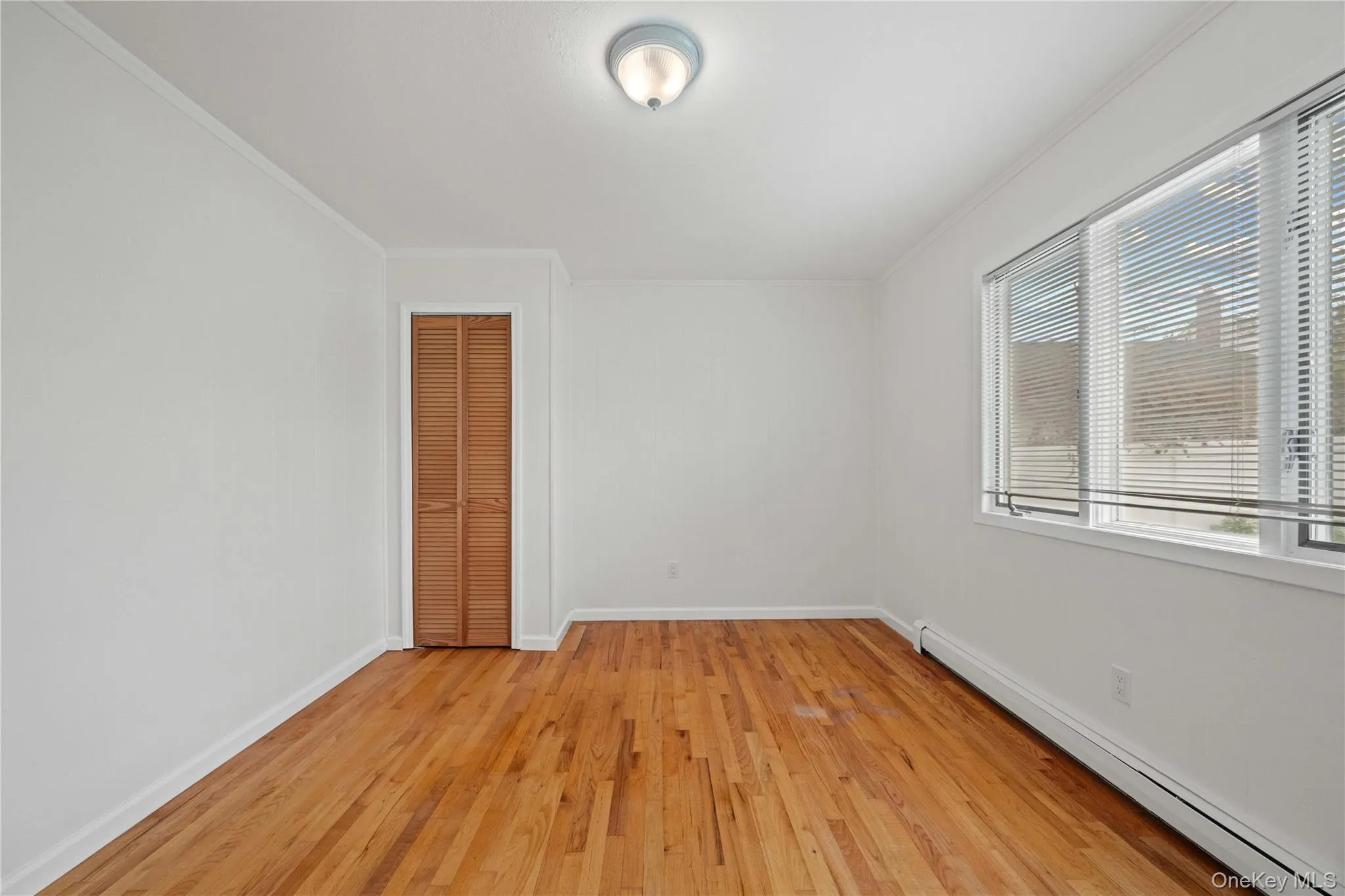 Hardwood flooring, painted walls, louvered closet doors, window with blinds, and ceiling-mounted light fixture Hardwood flooring, painted walls, louvered closet doors, window with blinds, and ceiling-mounted light fixture