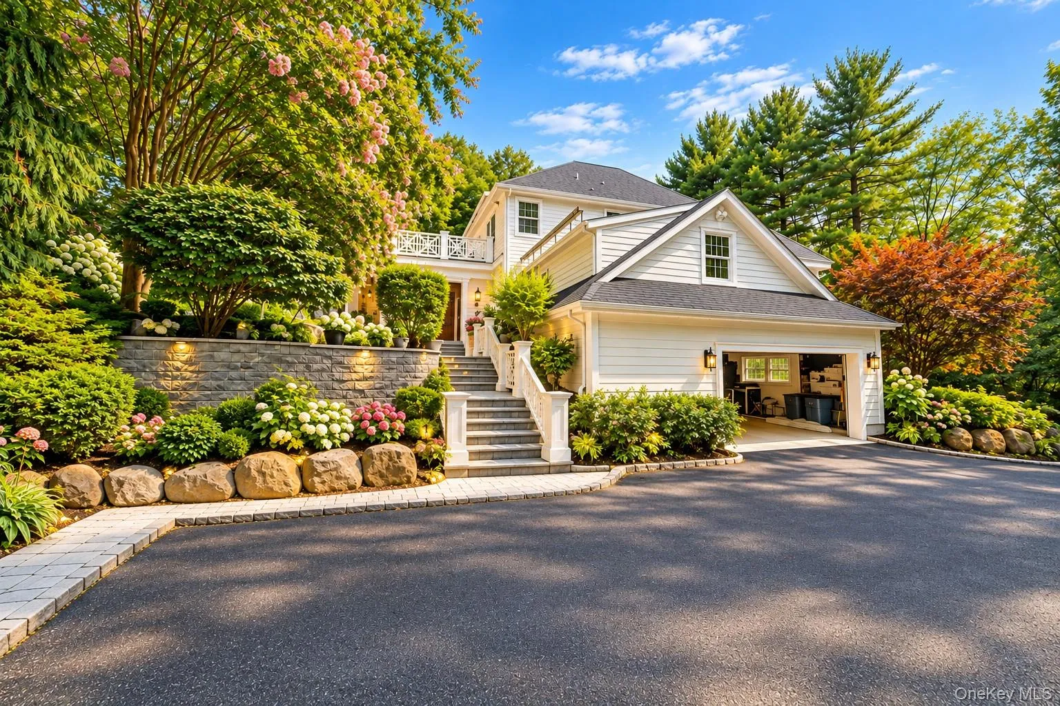White siding exterior with a dark shingle roof, attached garage, stone retaining walls, stone steps, and mature landscaping White siding exterior with a dark shingle roof, attached garage, stone retaining walls, stone steps, and mature landscaping