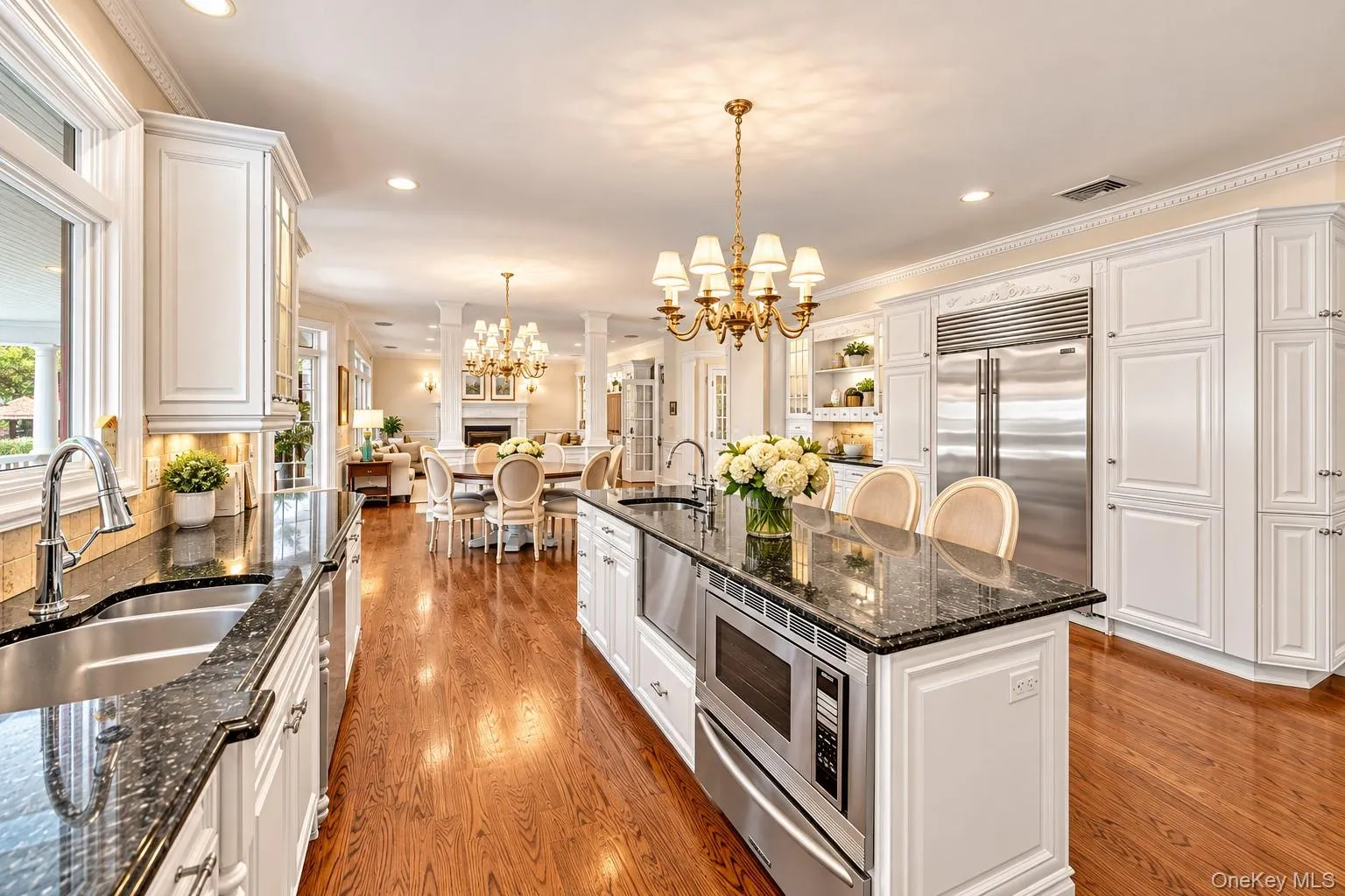 Chef's kitchen featuring white cabinetry with quartz countertops, a large center island with seating, stainless steel appliances including dual ovens and a gas cooktop, hardwood flooring throughout, recessed and chandelier lighting, and abundant natural light from multiple windows and glass doors Chef's kitchen featuring white cabinetry with quartz countertops, a large center island with seating, stainless steel appliances including dual ovens and a gas cooktop, hardwood flooring throughout, recessed and chandelier lighting, and abundant natural light from multiple windows and glass doors