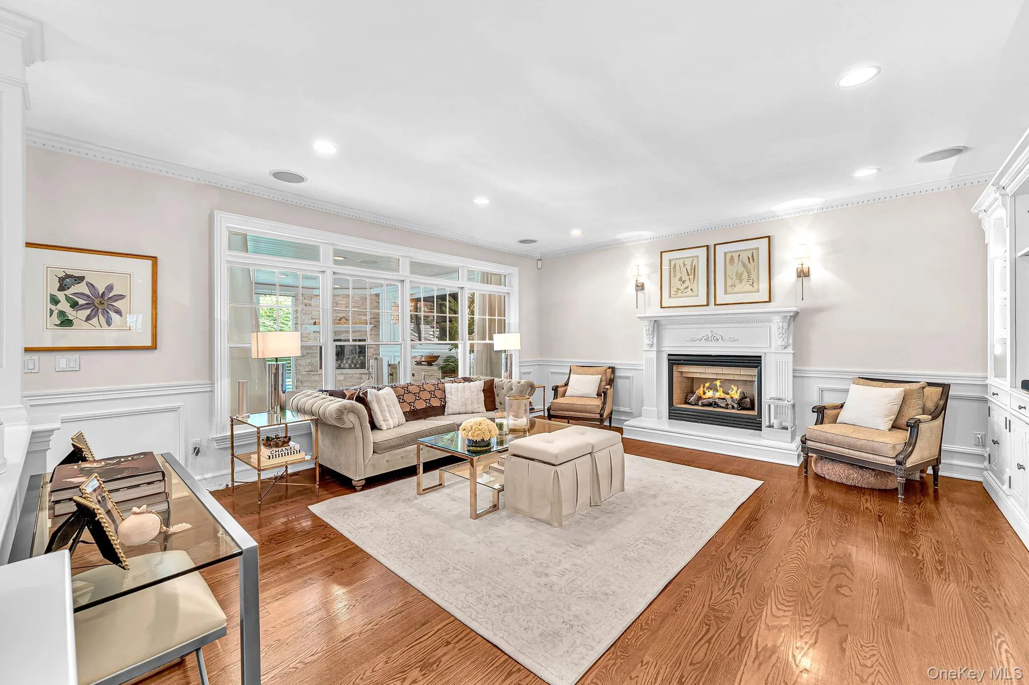 Elegant living room featuring hardwood flooring, a gas fireplace with decorative surround and mantel, recessed ceiling lighting, large multi-pane windows with natural light, and a neutral color palette with coordinating built-in wall treatments and wainscoting Elegant living room featuring hardwood flooring, a gas fireplace with decorative surround and mantel, recessed ceiling lighting, large multi-pane windows with natural light, and a neutral color palette with coordinating built-in wall treatments and wainscoting