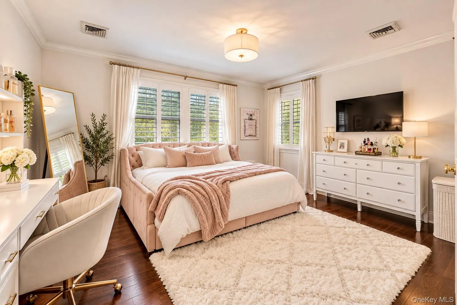 Spacious primary bedroom featuring multiple windows with plantation shutters and natural light, hardwood flooring, an ensuite bathroom, a walk-in closet, and a white wood dresser with integrated television mounting Spacious primary bedroom featuring multiple windows with plantation shutters and natural light, hardwood flooring, an ensuite bathroom, a walk-in closet, and a white wood dresser with integrated television mounting