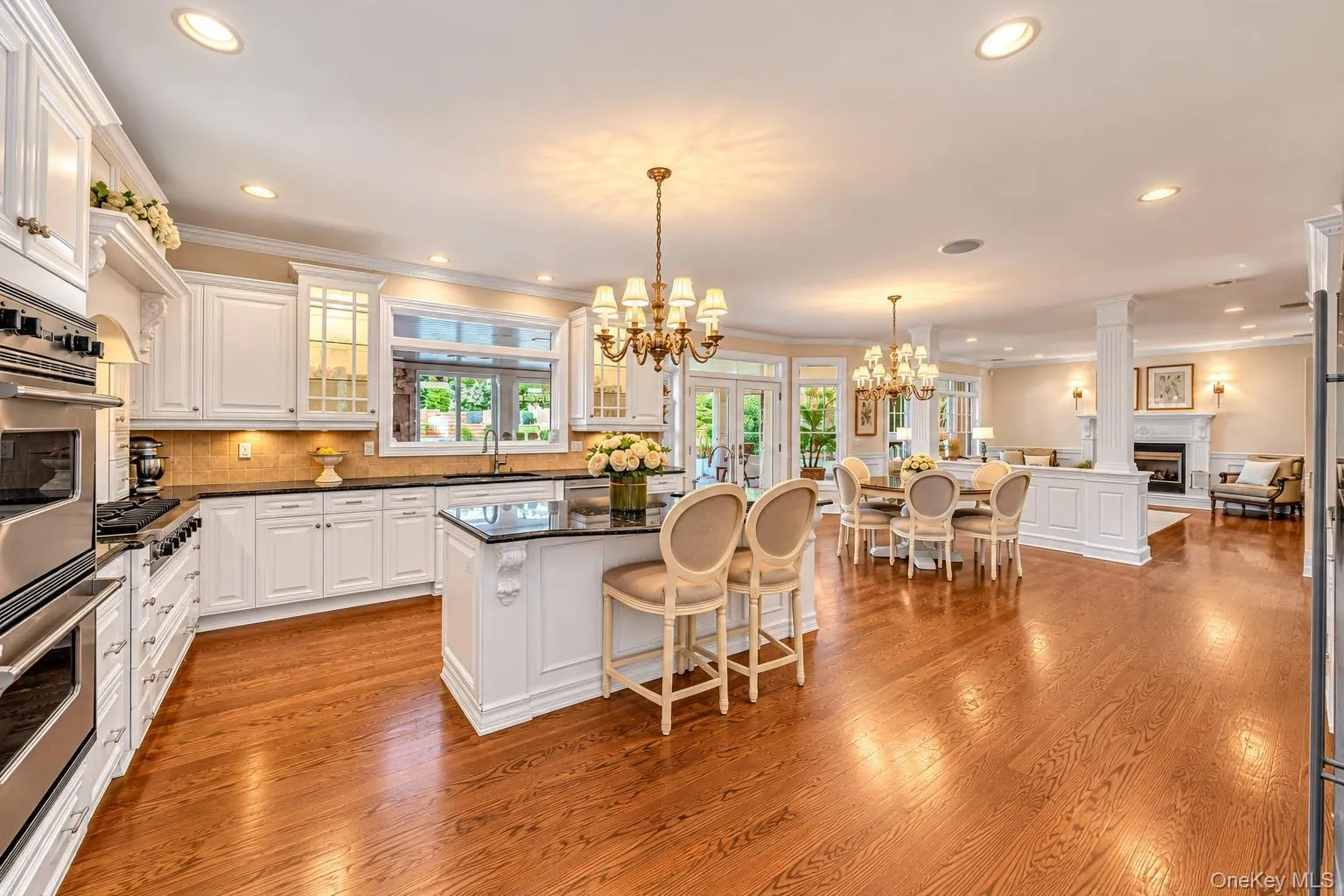 Gourmet kitchen featuring white cabinetry with glass-front doors, stainless steel double wall ovens, a large island with seating, quartz countertops, recessed and pendant lighting, and hardwood flooring that extends into the adjoining dining and living areas Gourmet kitchen featuring white cabinetry with glass-front doors, stainless steel double wall ovens, a large island with seating, quartz countertops, recessed and pendant lighting, and hardwood flooring that extends into the adjoining dining and living areas