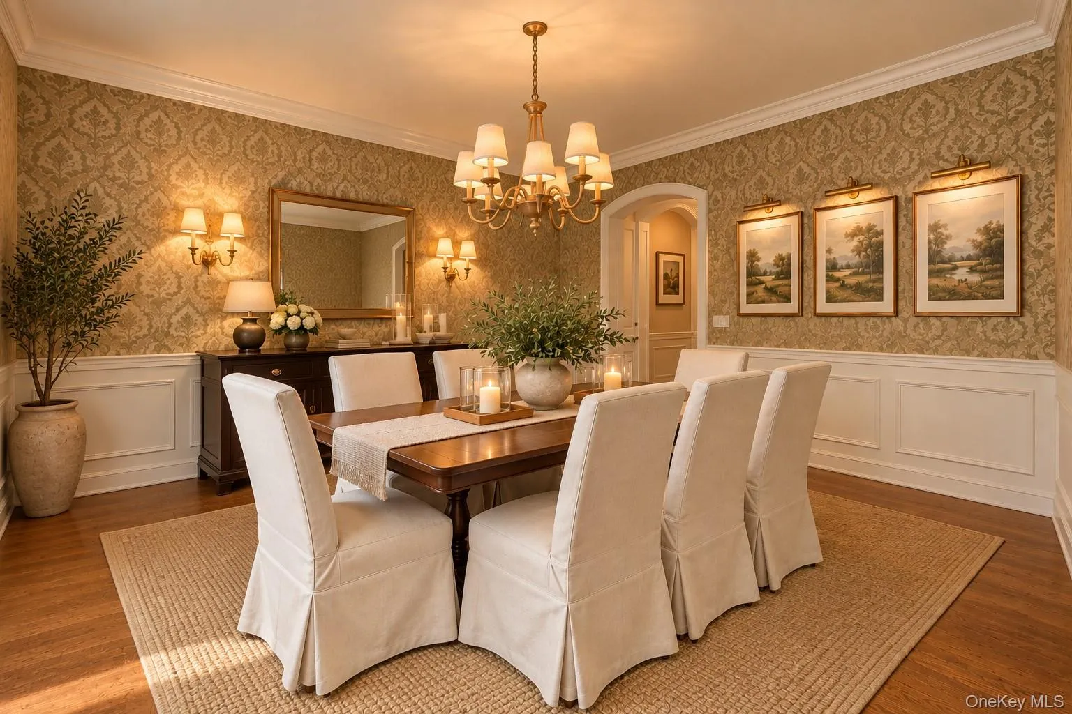Elegant formal dining room showcasing damask-patterned wallcovering, wainscoting with crown molding, hardwood flooring, and a multi-arm brass chandelier Elegant formal dining room showcasing damask-patterned wallcovering, wainscoting with crown molding, hardwood flooring, and a multi-arm brass chandelier