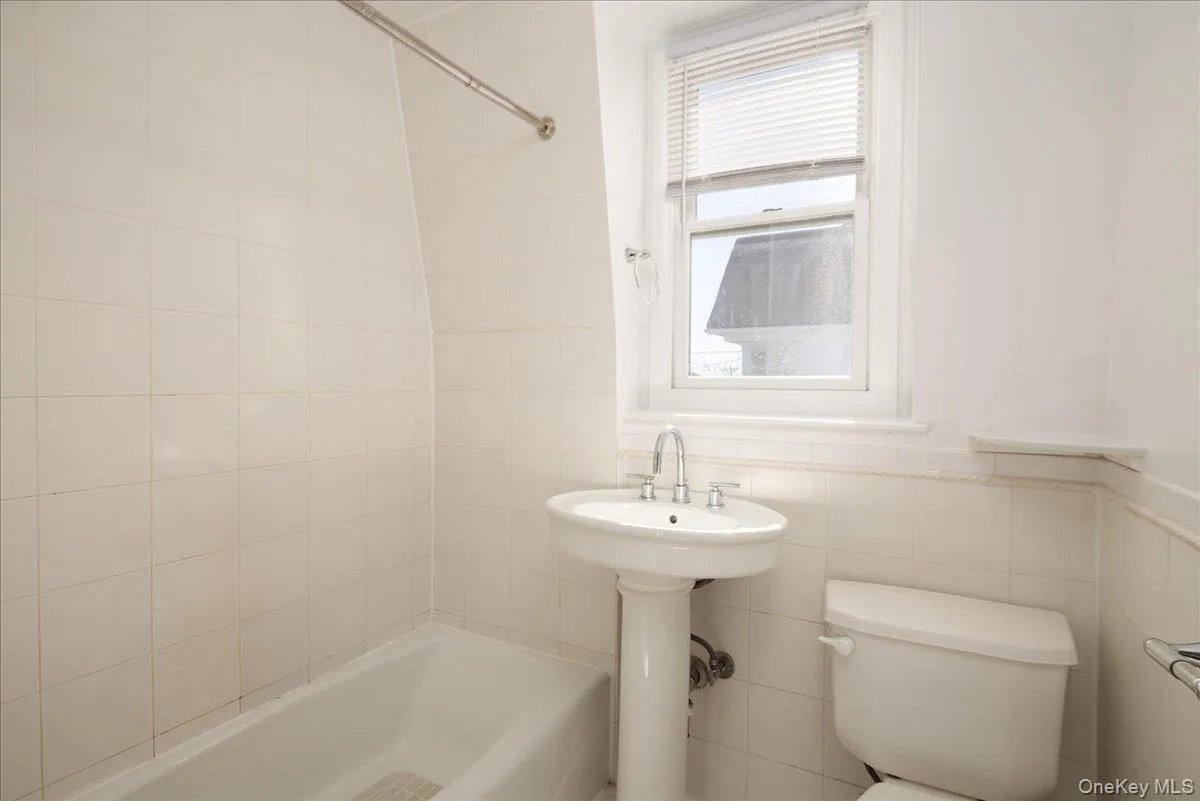 Second Floor Bathroom featuring ceramic wall tiling, a pedestal sink with a dual-handle chrome faucet, a recessed bathtub, a window with horizontal blinds, and a built-in corner shelf Second Floor Bathroom featuring ceramic wall tiling, a pedestal sink with a dual-handle chrome faucet, a recessed bathtub, a window with horizontal blinds, and a built-in corner shelf