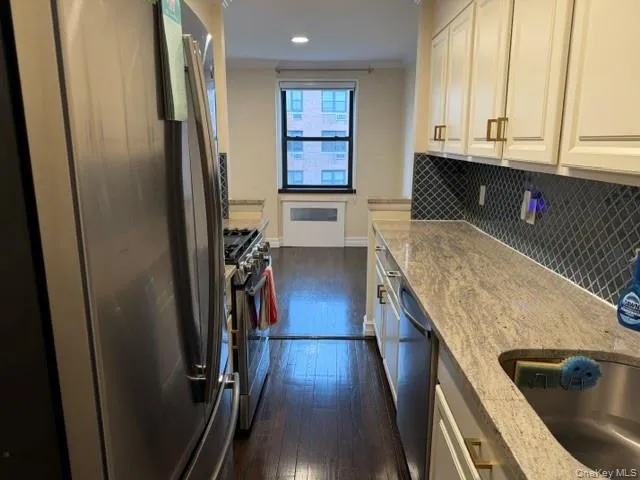 Galley kitchen featuring stainless steel appliances, white cabinetry with brass hardware, patterned tile backsplash, and light-toned countertops Galley kitchen featuring stainless steel appliances, white cabinetry with brass hardware, patterned tile backsplash, and light-toned countertops