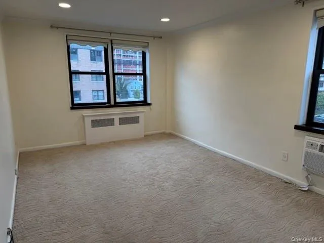 Recessed lighting, light-toned carpeting, two dark-framed windows, and a window-mounted air conditioning unit Recessed lighting, light-toned carpeting, two dark-framed windows, and a window-mounted air conditioning unit