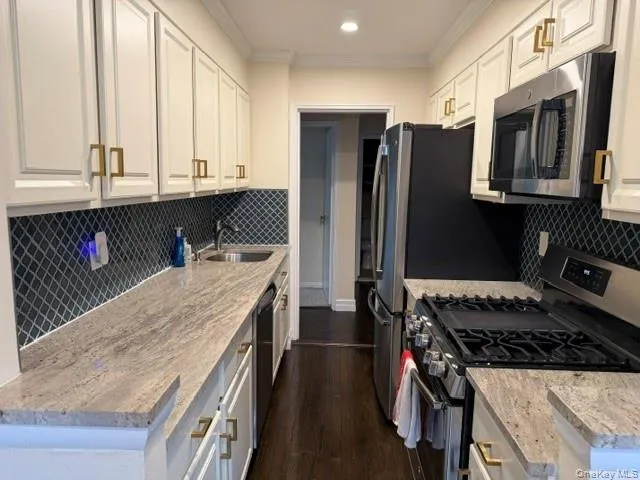 Galley kitchen featuring white cabinetry with gold-toned hardware, full-height decorative tile backsplash, light-toned stone countertops, and stainless steel appliances Galley kitchen featuring white cabinetry with gold-toned hardware, full-height decorative tile backsplash, light-toned stone countertops, and stainless steel appliances