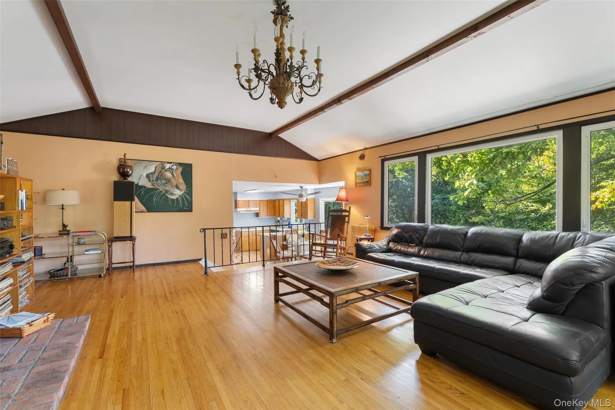 Vaulted ceiling with exposed wood beams, hardwood flooring, expansive window wall, brick fireplace, and a chandelier light fixture. Vaulted ceiling with exposed wood beams, hardwood flooring, expansive window wall, brick fireplace, and a chandelier light fixture.