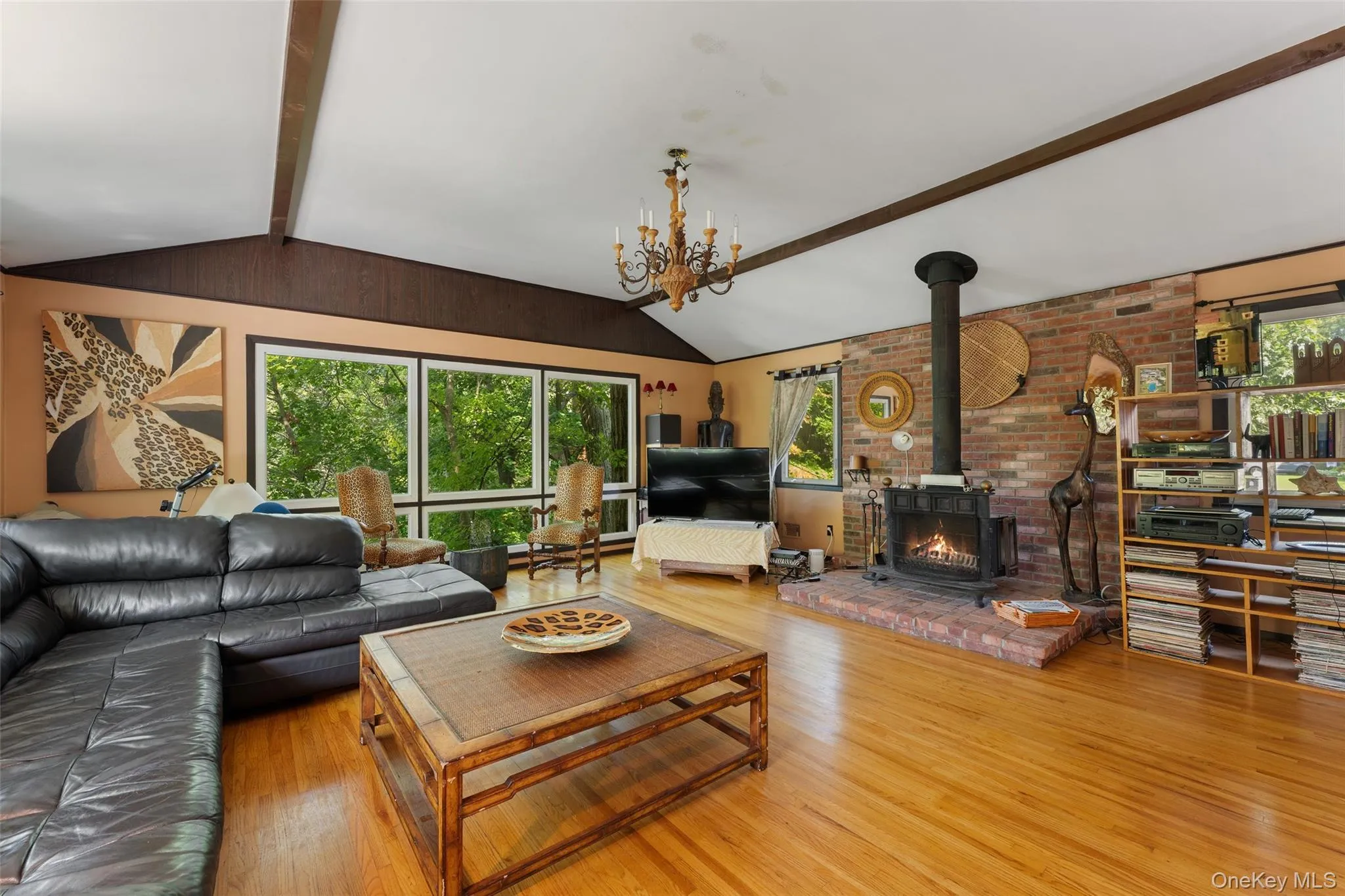 Vaulted ceiling with exposed wood beams, hardwood flooring, expansive window wall, brick fireplace, and a chandelier light fixture. Vaulted ceiling with exposed wood beams, hardwood flooring, expansive window wall, brick fireplace, and a chandelier light fixture.