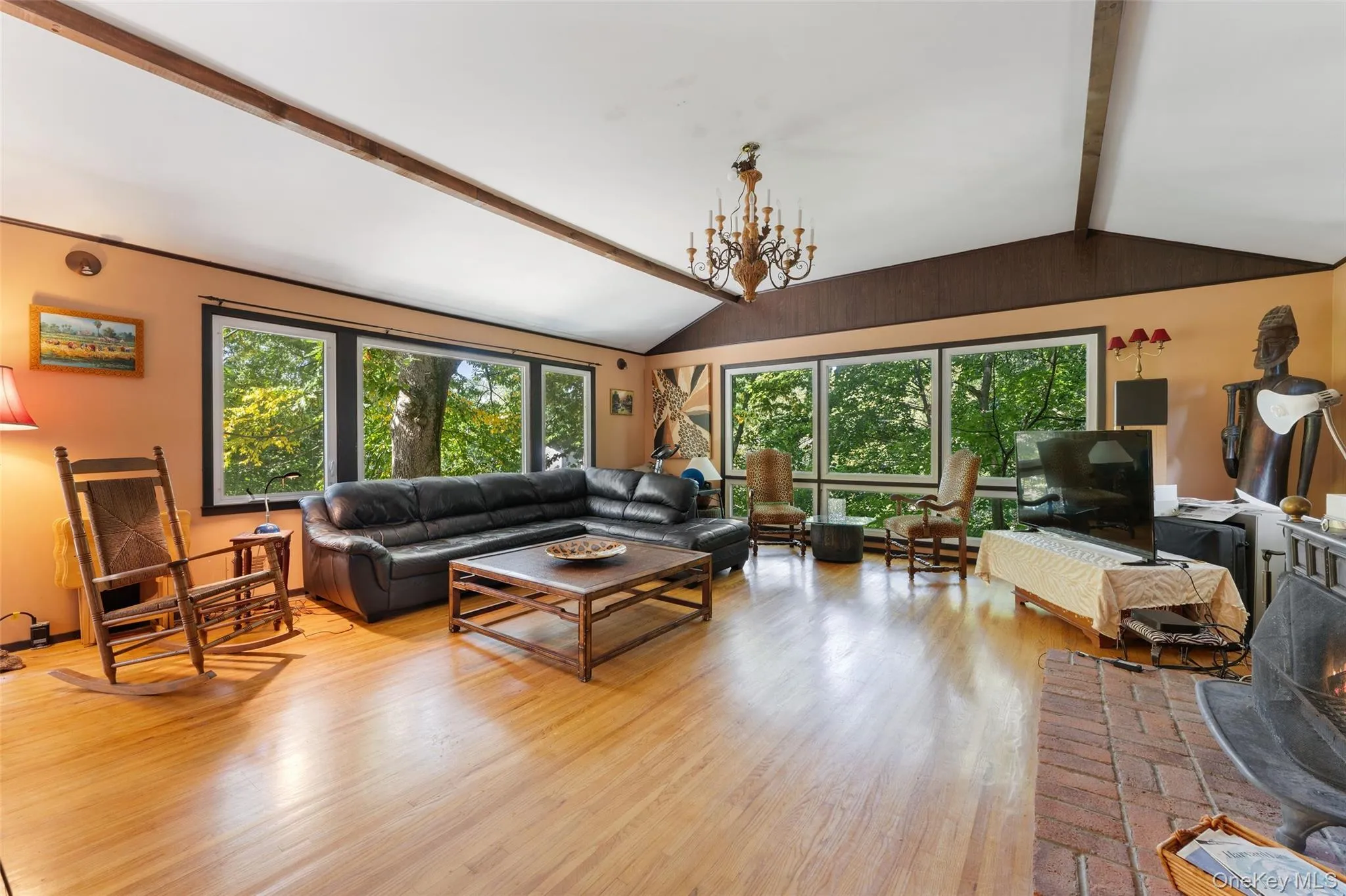 Vaulted ceiling with exposed wood beams, hardwood flooring, expansive window wall, brick fireplace, and a chandelier light fixture. Vaulted ceiling with exposed wood beams, hardwood flooring, expansive window wall, brick fireplace, and a chandelier light fixture.
