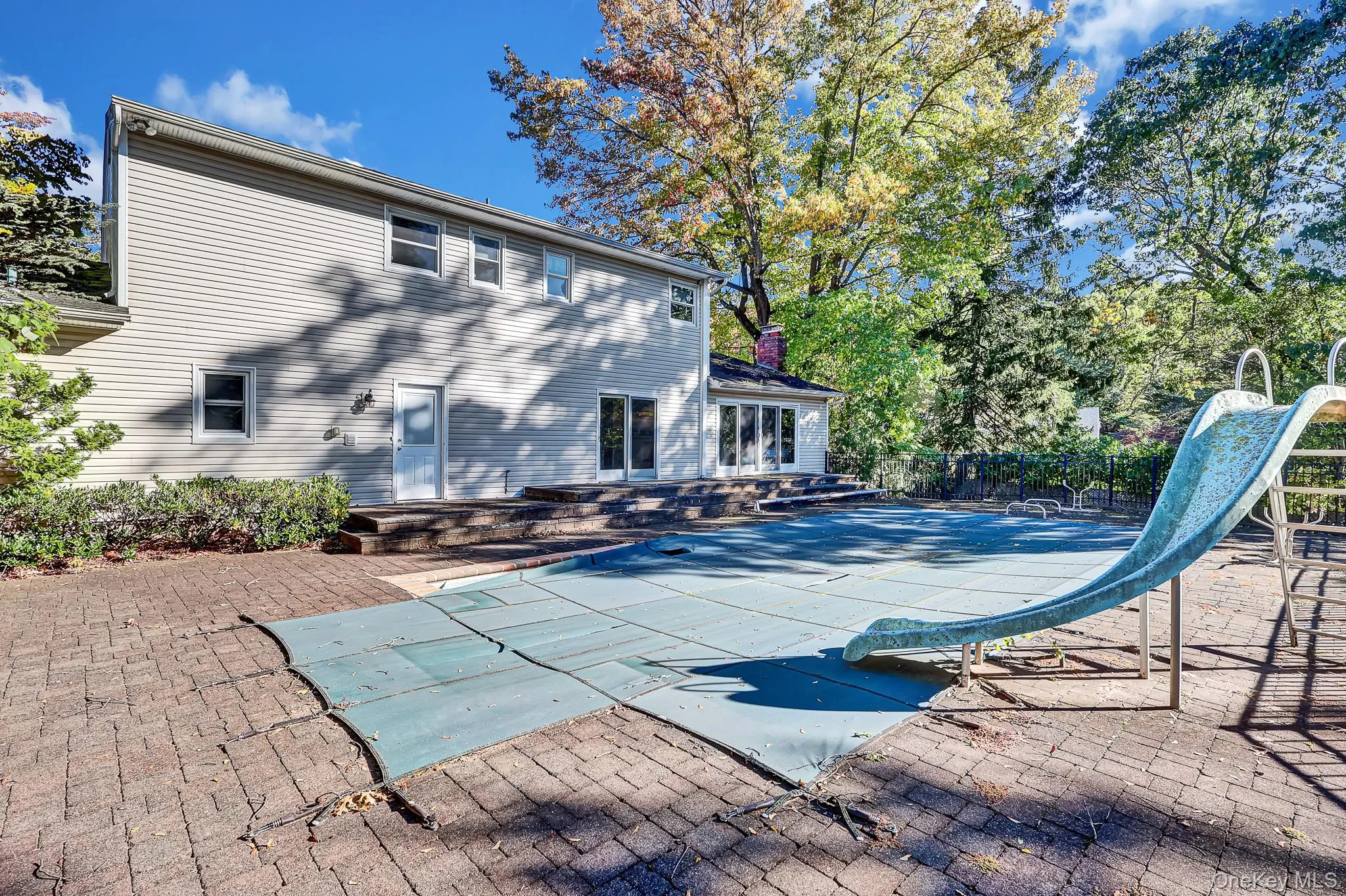 Exterior view of the residence featuring horizontal siding, a multi-level stone paver patio, an in-ground pool with a slide, and mature trees Exterior view of the residence featuring horizontal siding, a multi-level stone paver patio, an in-ground pool with a slide, and mature trees