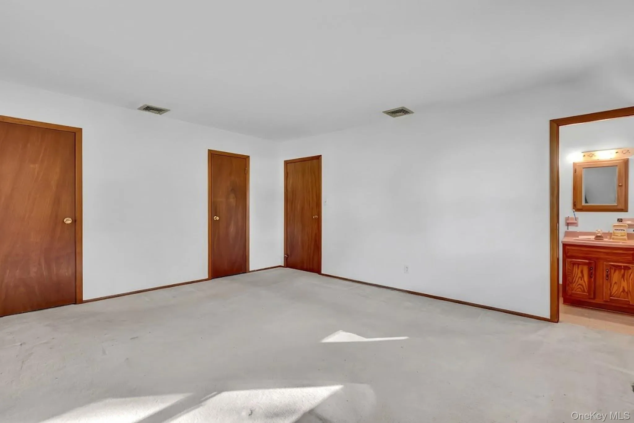 Spacious room featuring neutral carpeting, three solid wood interior doors, and an entry to an attached bathroom with a wood vanity and vanity mirror Spacious room featuring neutral carpeting, three solid wood interior doors, and an entry to an attached bathroom with a wood vanity and vanity mirror