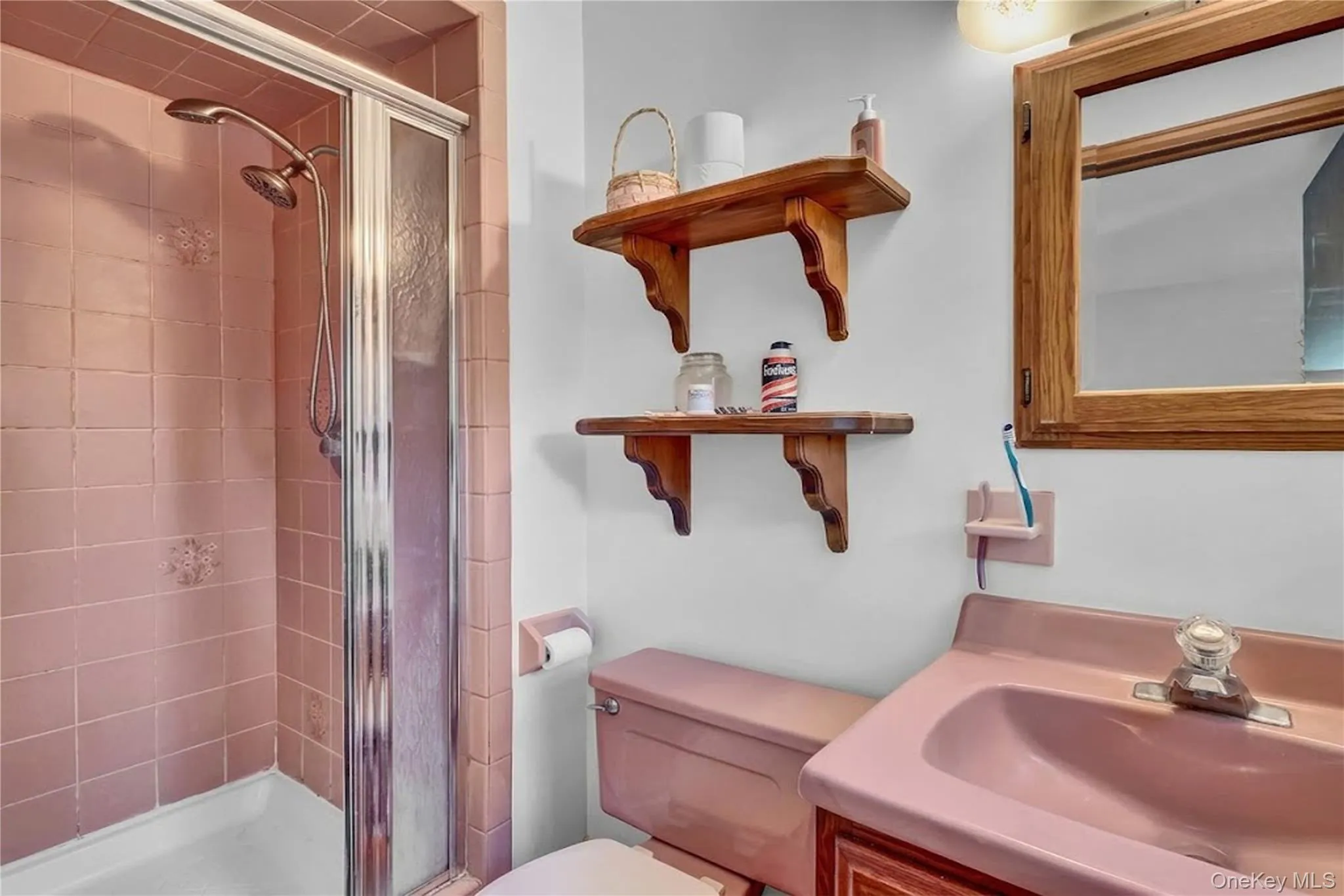 Walk-in shower with metal-framed glass enclosure, pink tile surround, integrated pink vanity with basin, wooden medicine cabinet, and wall-mounted wooden shelving Walk-in shower with metal-framed glass enclosure, pink tile surround, integrated pink vanity with basin, wooden medicine cabinet, and wall-mounted wooden shelving