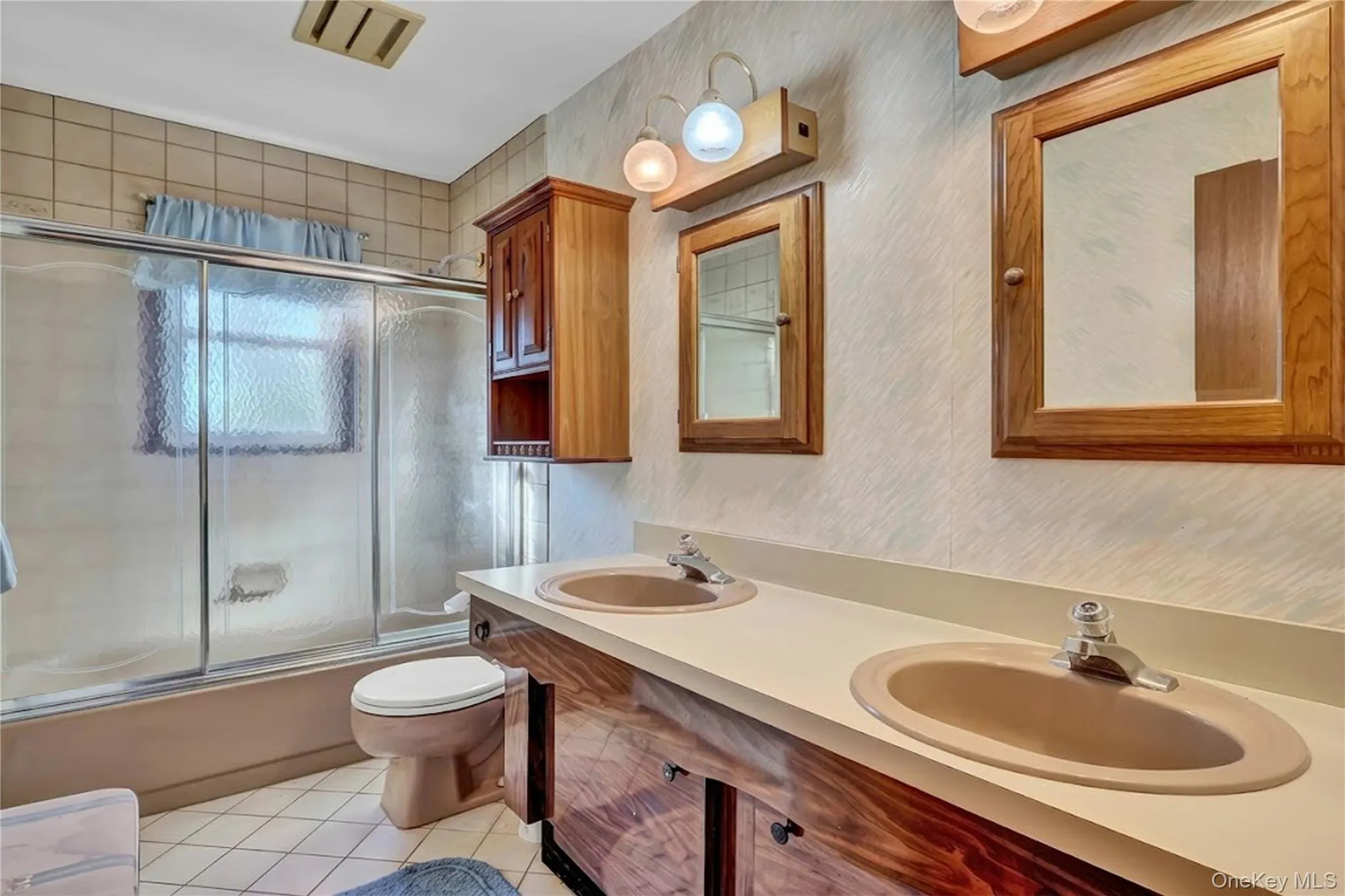 Bathroom featuring a double-sink vanity with wood-finish cabinetry, integrated countertop with dual basins, a glass-enclosed shower and tub combination, and wood-framed medicine cabinets Bathroom featuring a double-sink vanity with wood-finish cabinetry, integrated countertop with dual basins, a glass-enclosed shower and tub combination, and wood-framed medicine cabinets