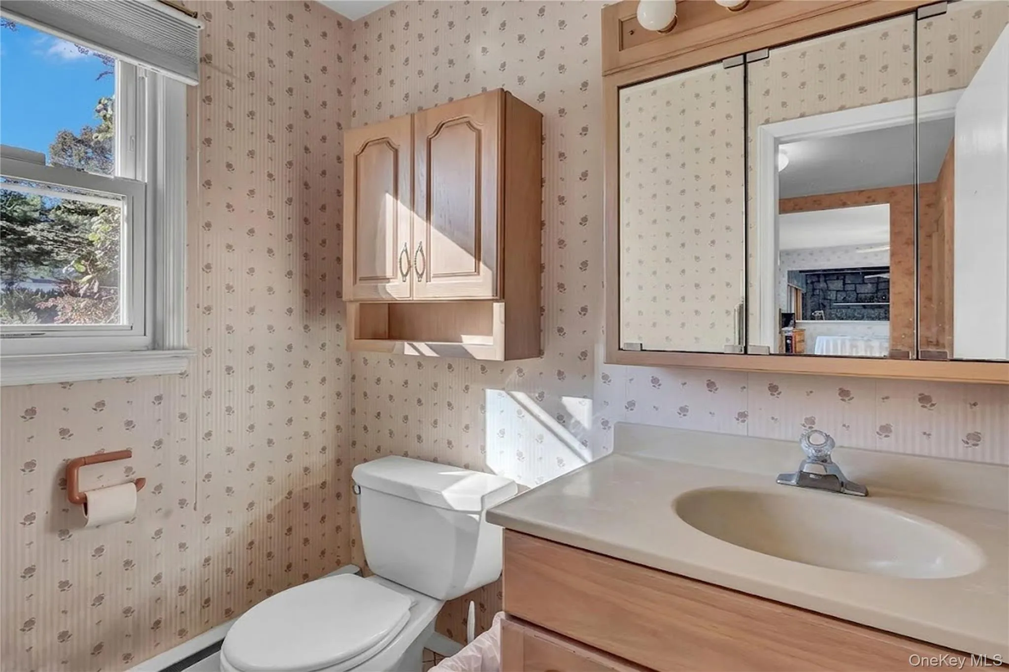 Bathroom featuring wood-finish cabinetry, a built-in medicine cabinet with integrated lighting, a single-basin vanity, and a window with a valance Bathroom featuring wood-finish cabinetry, a built-in medicine cabinet with integrated lighting, a single-basin vanity, and a window with a valance