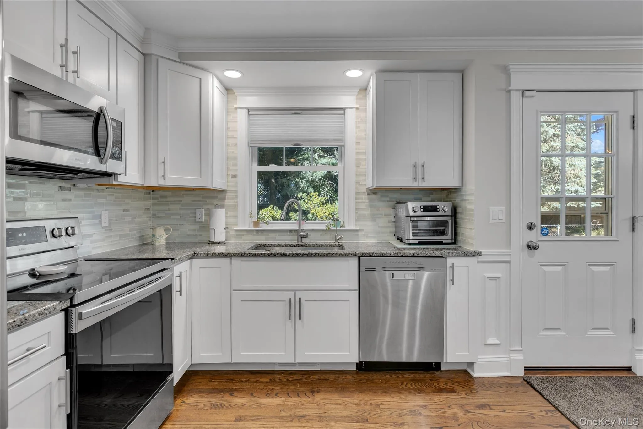 Kitchen featuring white cabinetry, stainless steel appliances, granite countertops, a tiled backsplash, and wood-finish flooring, access to backyard rear patio Kitchen featuring white cabinetry, stainless steel appliances, granite countertops, a tiled backsplash, and wood-finish flooring, access to backyard rear patio