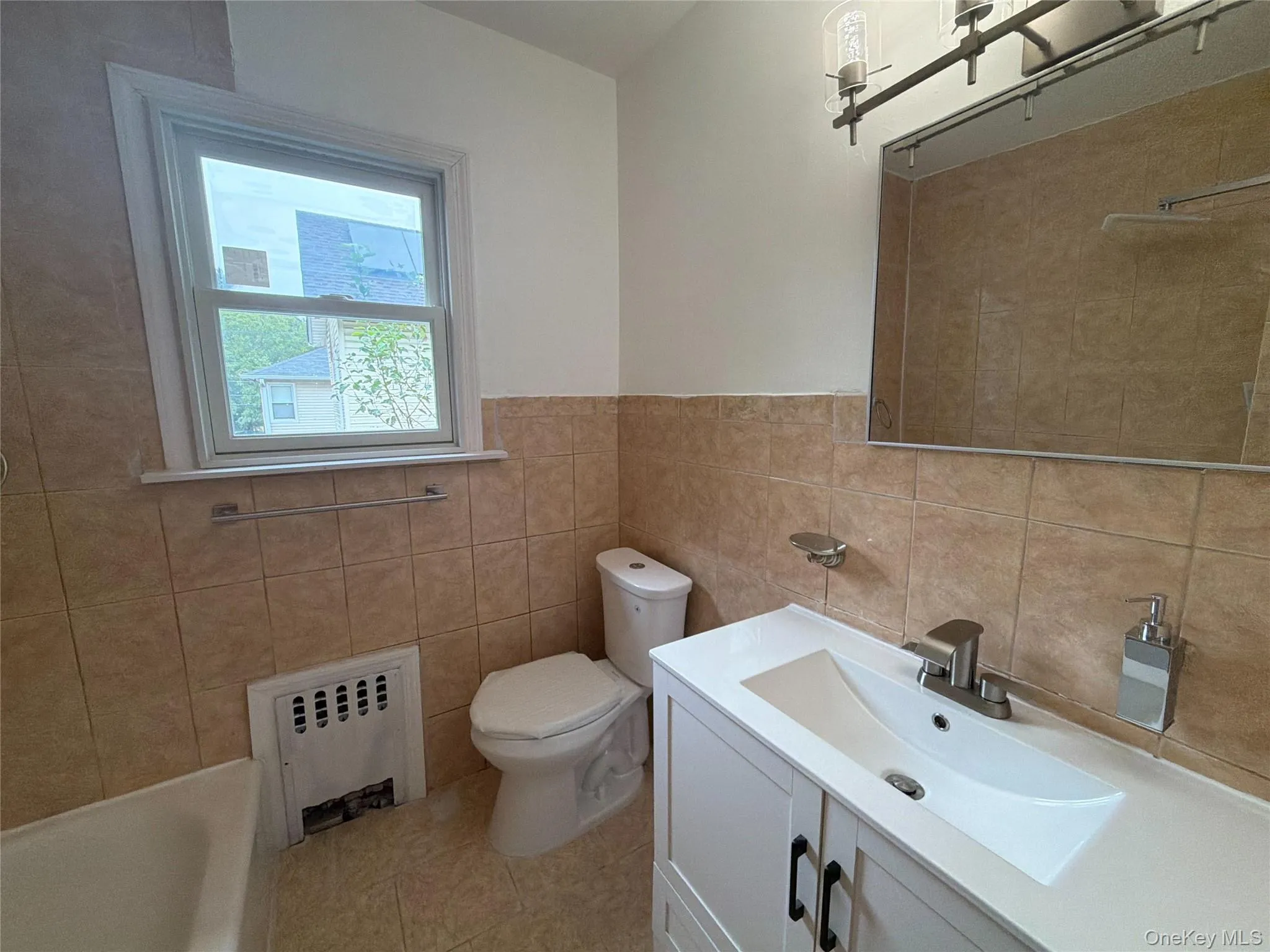 Tiled bathroom featuring a white vanity with integrated sink, brushed nickel faucet, large wall mirror, and a window with trim Tiled bathroom featuring a white vanity with integrated sink, brushed nickel faucet, large wall mirror, and a window with trim