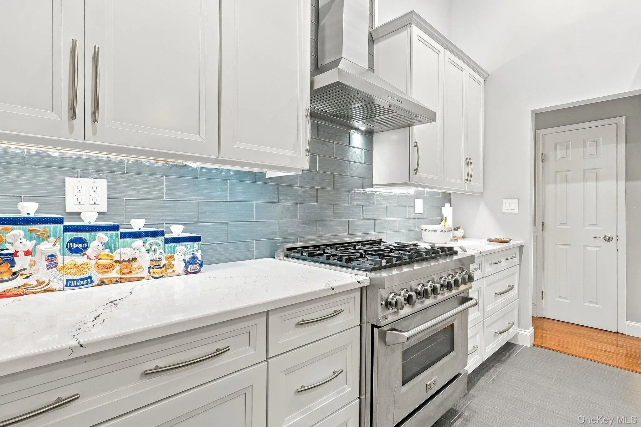 White cabinetry with brushed nickel hardware, quartz countertops, a stainless-steel range with a ventilation hood, and a light blue tile backsplash White cabinetry with brushed nickel hardware, quartz countertops, a stainless-steel range with a ventilation hood, and a light blue tile backsplash