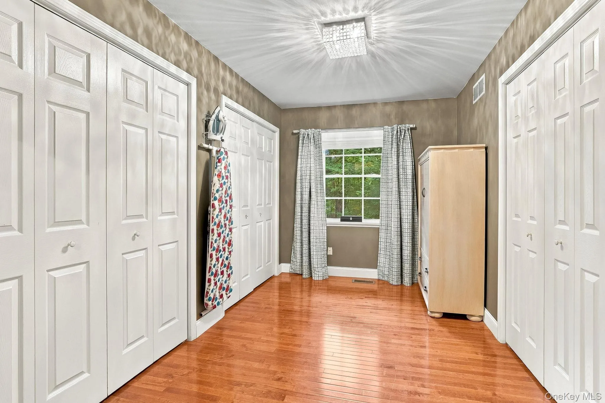 Hardwood-flooring, multiple paneled doors, single window, and a ceiling-mounted light fixture-Primary Closet Hardwood-flooring, multiple paneled doors, single window, and a ceiling-mounted light fixture-Primary Closet