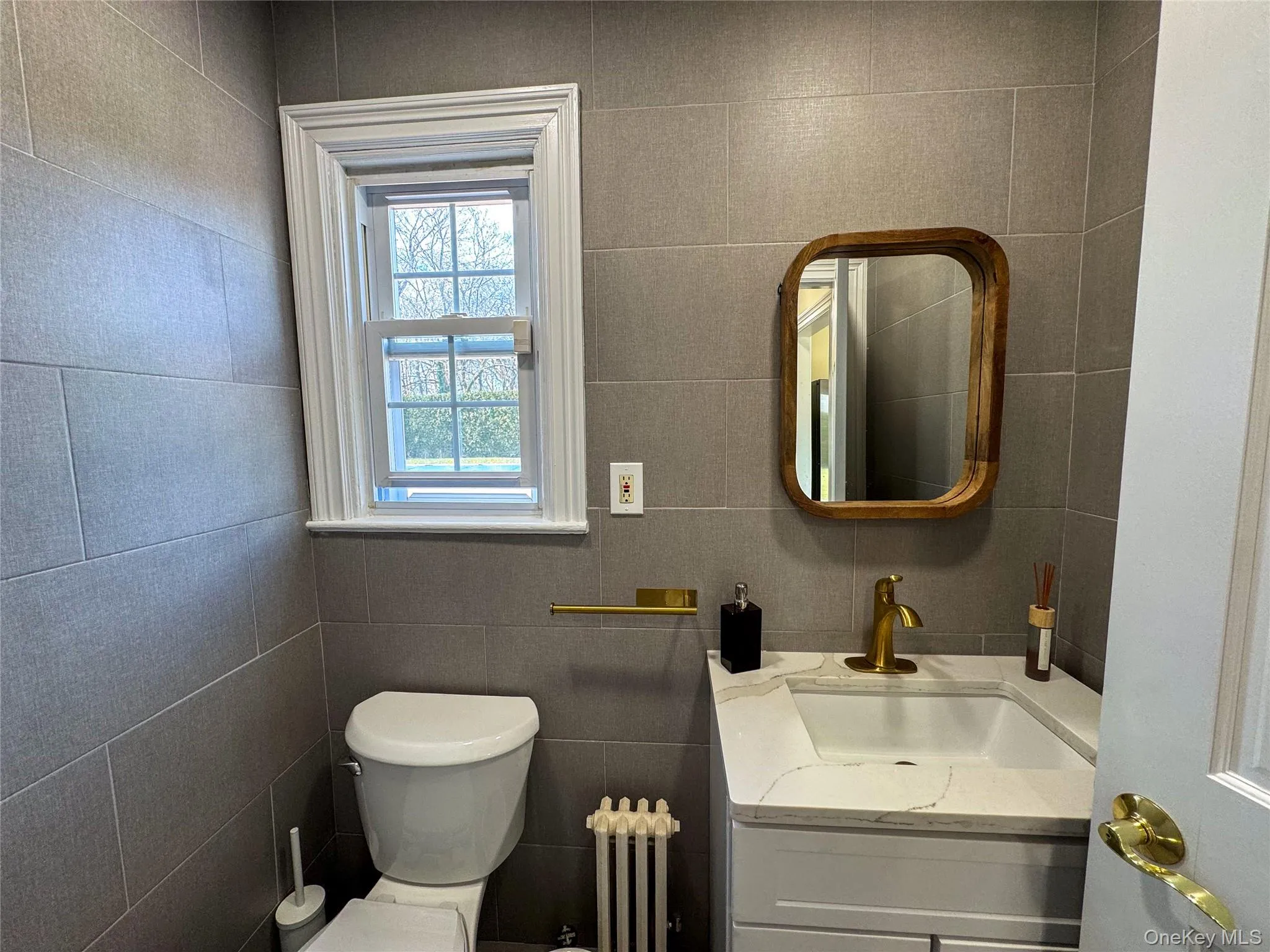 Gray tile walls, single hung window with white trim, white toilet, white vanity with integrated sink, brass-finish faucet Gray tile walls, single hung window with white trim, white toilet, white vanity with integrated sink, brass-finish faucet