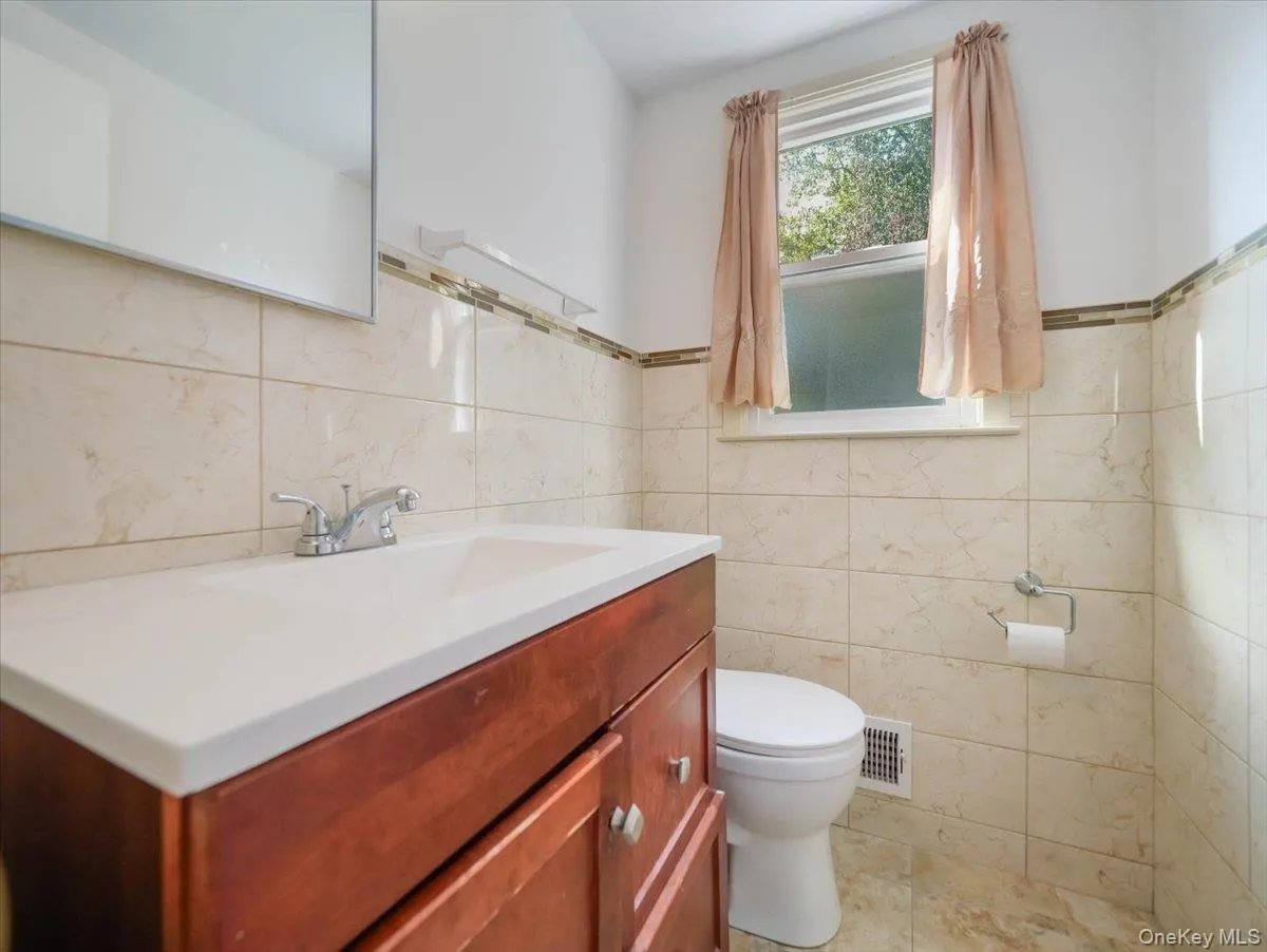 Bathroom featuring a wood-finish vanity with a white integrated sink, full-height wall tiling with a mosaic accent border, and a large wall-mounted mirror Bathroom featuring a wood-finish vanity with a white integrated sink, full-height wall tiling with a mosaic accent border, and a large wall-mounted mirror