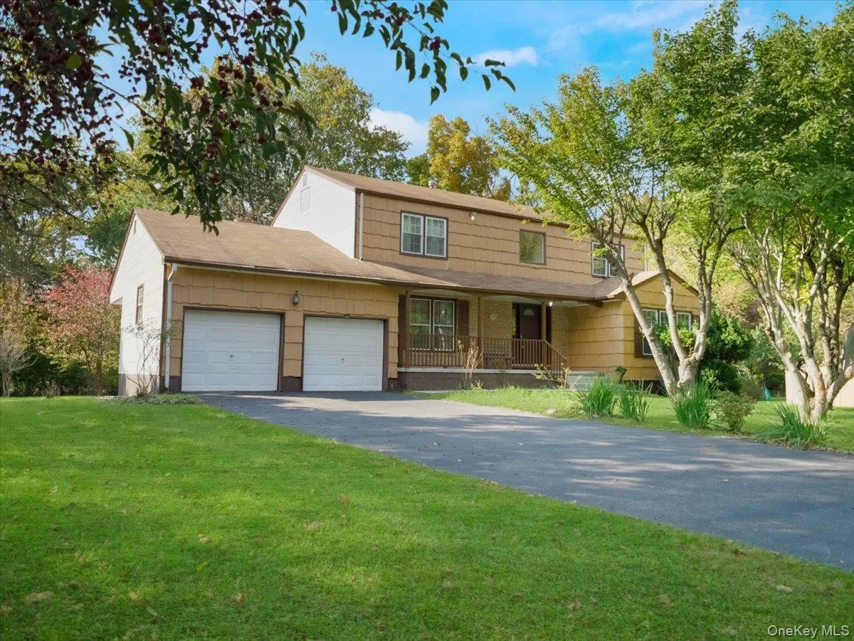 Two-story exterior featuring tan horizontal siding, a covered front porch with railing, an attached two-car garage, and an asphalt driveway situated on a landscaped lot with mature trees Two-story exterior featuring tan horizontal siding, a covered front porch with railing, an attached two-car garage, and an asphalt driveway situated on a landscaped lot with mature trees