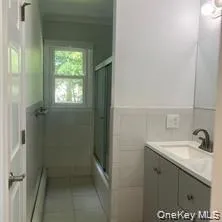 Bathroom featuring a window, white wall tiling, a shower-tub combination with sliding glass doors, a vanity with an integrated sink, and a wall-mounted mirror Bathroom featuring a window, white wall tiling, a shower-tub combination with sliding glass doors, a vanity with an integrated sink, and a wall-mounted mirror
