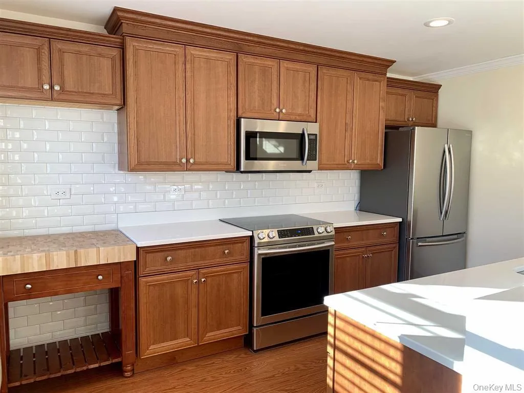 Kitchen with appliances with stainless steel finishes, ornamental molding, brown cabinetry, light wood-style flooring, and light countertops Kitchen with appliances with stainless steel finishes, ornamental molding, brown cabinetry, light wood-style flooring, and light countertops