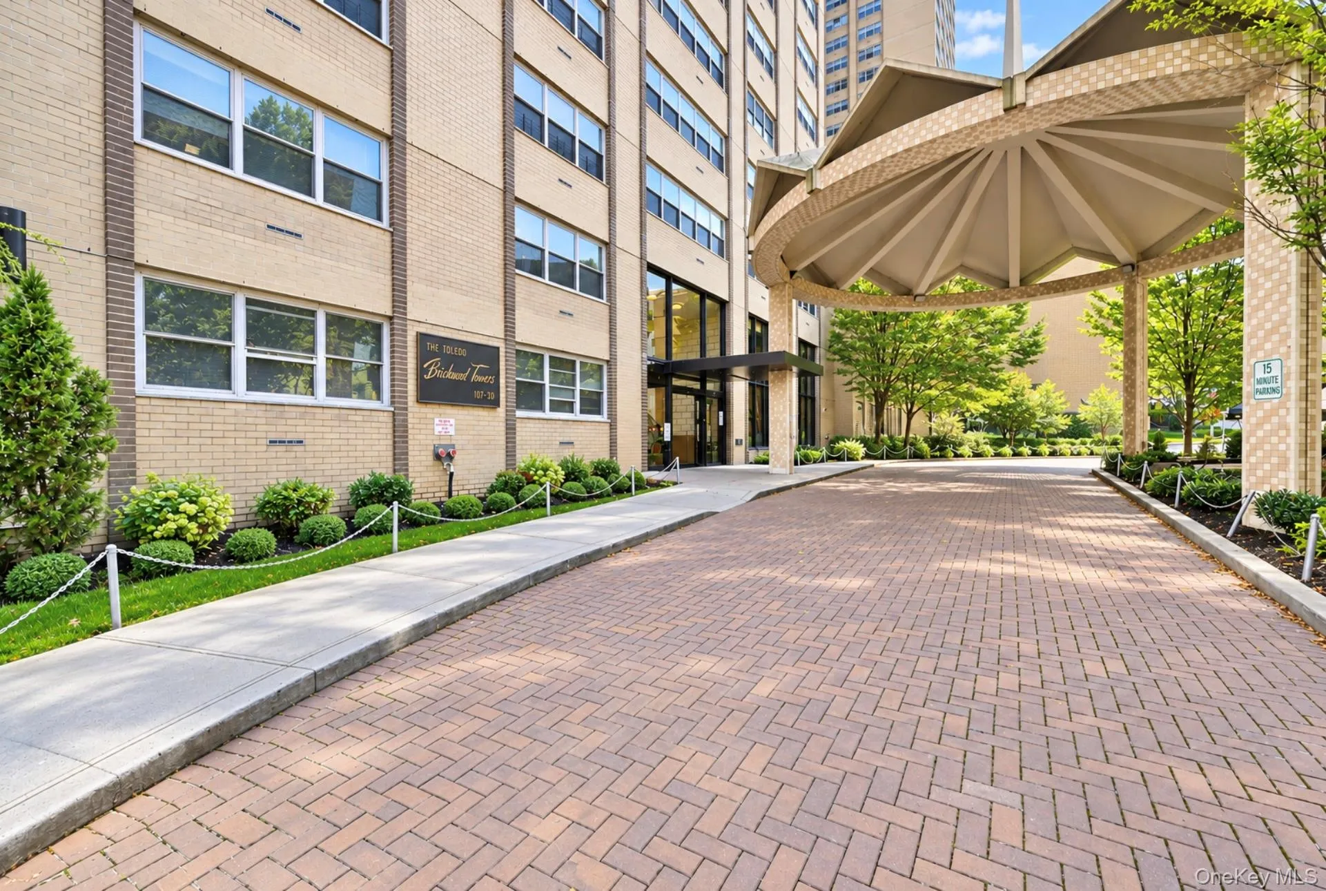 Brick-patterned driveway leading to a covered entrance with a multi-paneled canopy, featuring a brick facade building and structured landscaping Brick-patterned driveway leading to a covered entrance with a multi-paneled canopy, featuring a brick facade building and structured landscaping