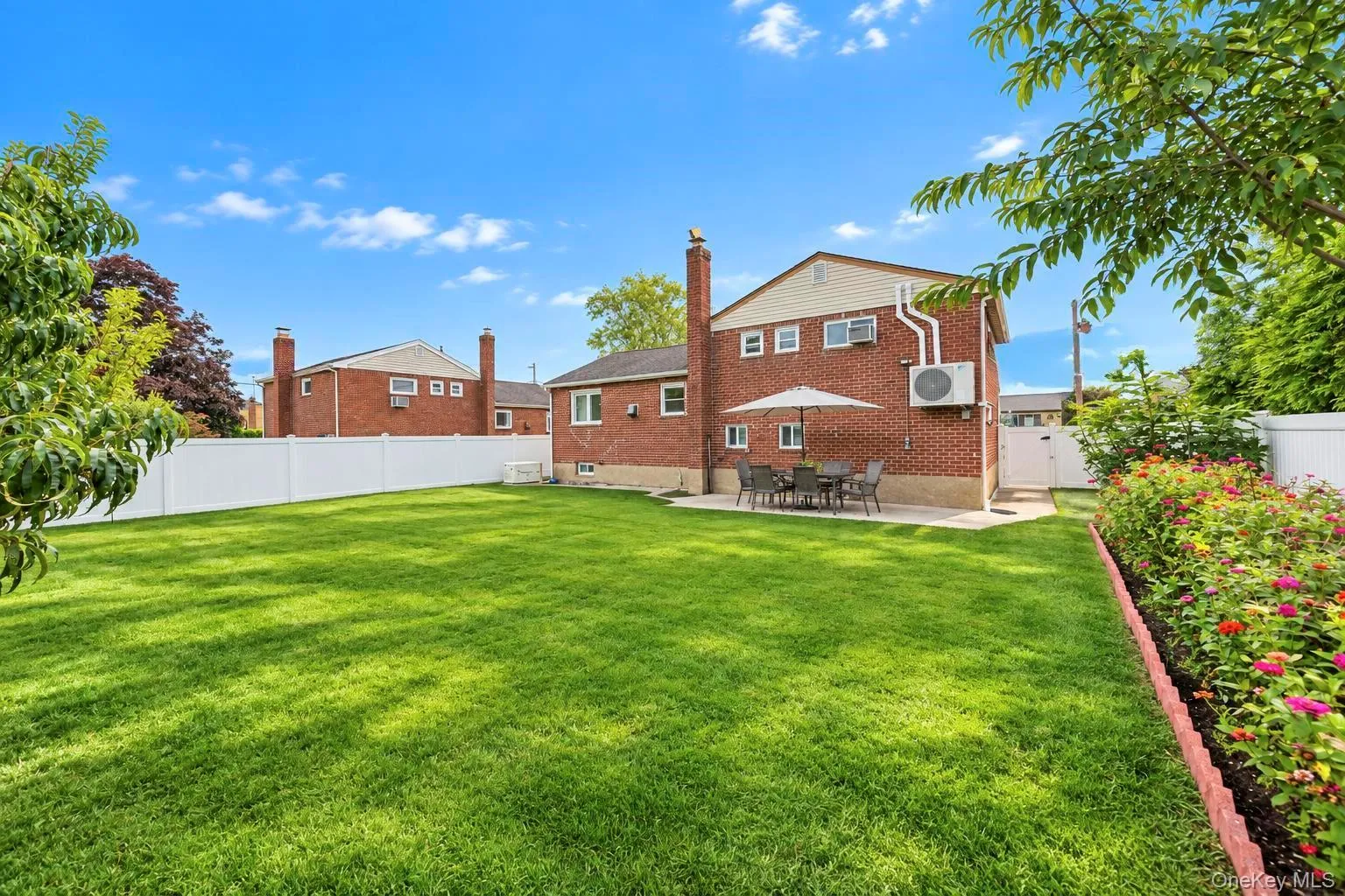 Expansive green lawn, brick exterior, white privacy fencing, concrete patio area, and a planted border with flowering shrubs Expansive green lawn, brick exterior, white privacy fencing, concrete patio area, and a planted border with flowering shrubs