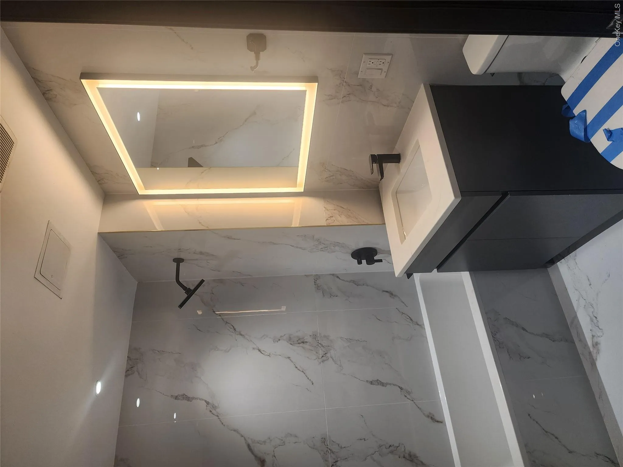 Bathroom featuring a tub with a wall-mounted showerhead, a vanity with an integrated sink, an illuminated mirror, and marble-patterned wall and floor tiling Bathroom featuring a tub with a wall-mounted showerhead, a vanity with an integrated sink, an illuminated mirror, and marble-patterned wall and floor tiling