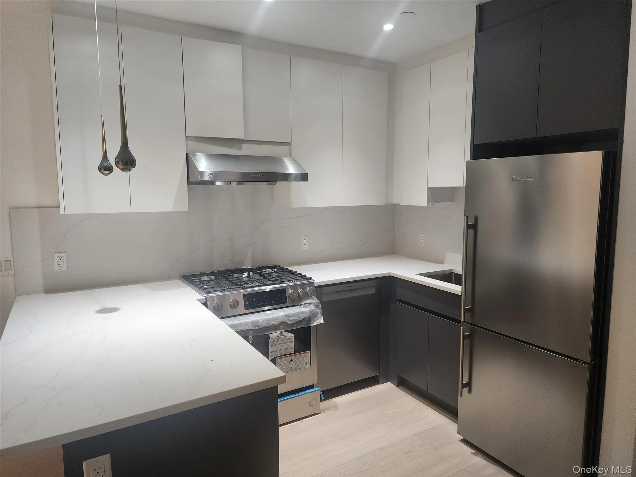 Modern kitchen featuring white upper cabinetry, dark lower cabinetry, stainless steel appliances, light-toned countertops, and light wood-finish flooring Modern kitchen featuring white upper cabinetry, dark lower cabinetry, stainless steel appliances, light-toned countertops, and light wood-finish flooring
