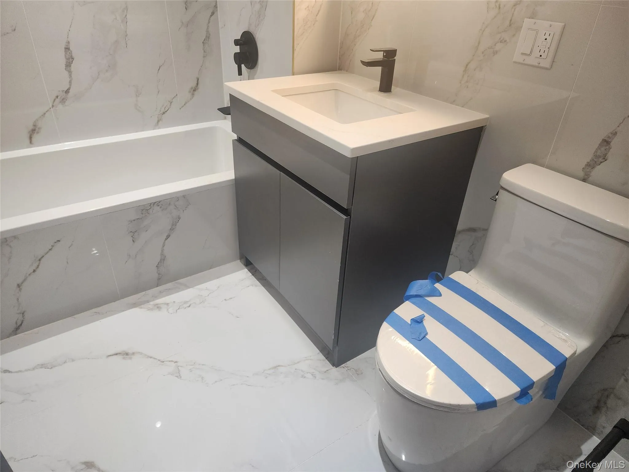 White tiled flooring with subtle veining, contemporary vanity with a white countertop and rectangular sink, and a white bathtub with tiled surround White tiled flooring with subtle veining, contemporary vanity with a white countertop and rectangular sink, and a white bathtub with tiled surround