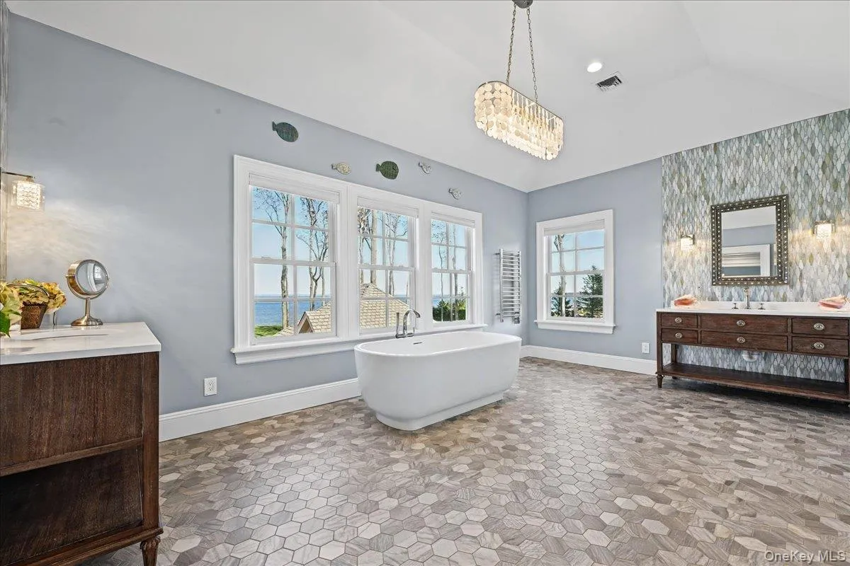 Hexagonal tile flooring, freestanding soaking tub, dual wood-finish vanity with countertop, textural accent wall, and multiple windows Hexagonal tile flooring, freestanding soaking tub, dual wood-finish vanity with countertop, textural accent wall, and multiple windows
