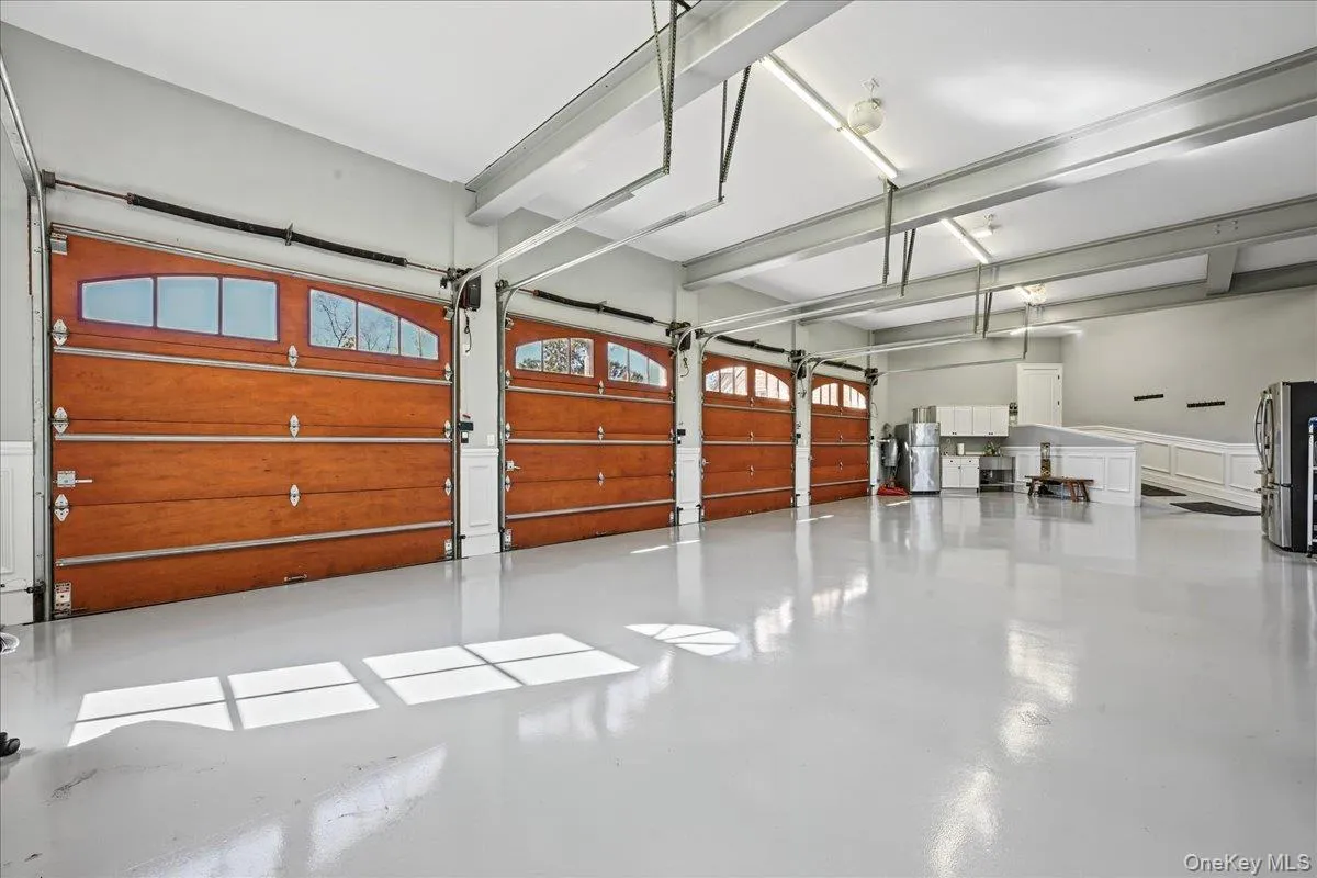 Multiple wood-finish garage doors with arched window panels, expansive light gray flooring, exposed overhead support beams, recessed lighting, and wall-mounted cabinetry Multiple wood-finish garage doors with arched window panels, expansive light gray flooring, exposed overhead support beams, recessed lighting, and wall-mounted cabinetry