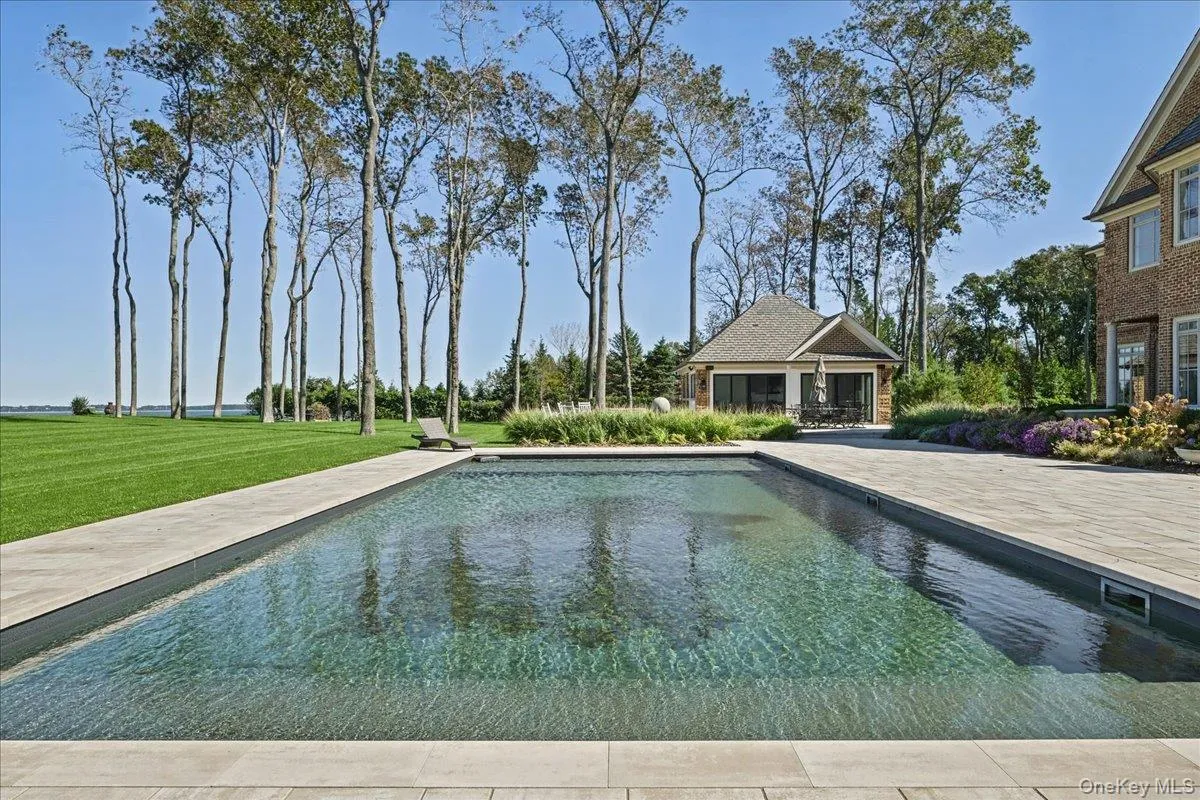 Expansive outdoor area featuring a swimming pool, a large patio, a separate pool house structure, a vast lawn, and mature trees Expansive outdoor area featuring a swimming pool, a large patio, a separate pool house structure, a vast lawn, and mature trees