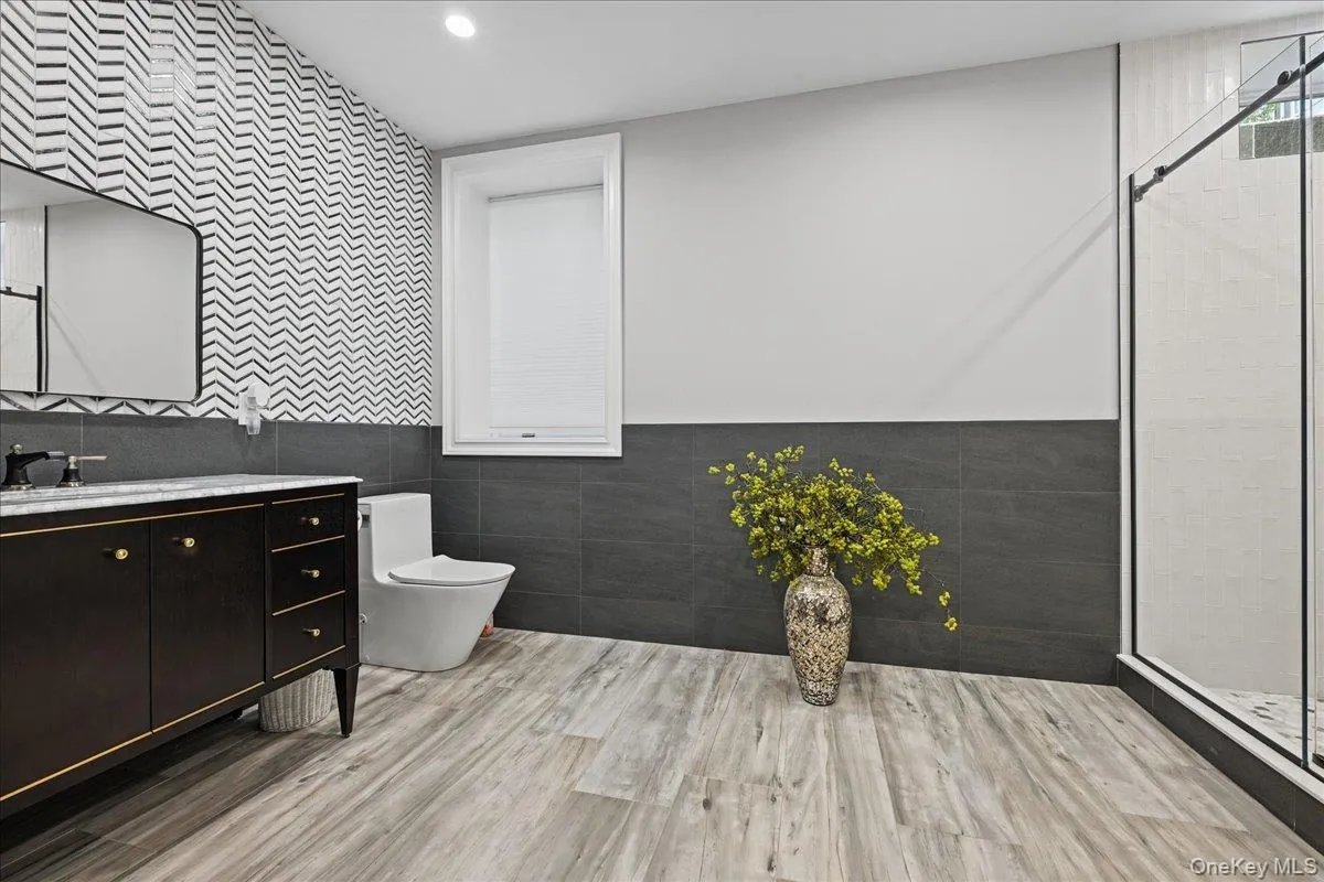 Modern bathroom featuring wood-finish flooring, a freestanding vanity with gold-tone hardware, a frameless shower enclosure, and a window with blinds Modern bathroom featuring wood-finish flooring, a freestanding vanity with gold-tone hardware, a frameless shower enclosure, and a window with blinds