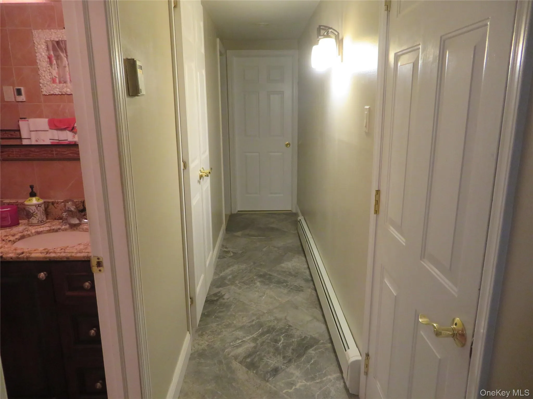 Hallway featuring stone-finish tile flooring, white paneled interior doors with brass-tone hardware, and wall-mounted lighting Hallway featuring stone-finish tile flooring, white paneled interior doors with brass-tone hardware, and wall-mounted lighting