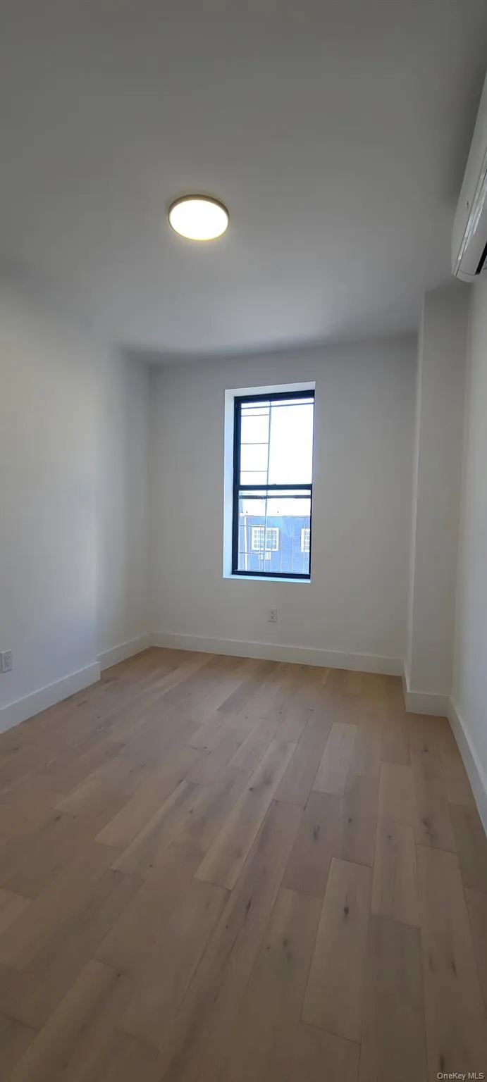 Hardwood-style flooring, single window with dark frame, flush-mount ceiling light, and wall-mounted climate control unit Hardwood-style flooring, single window with dark frame, flush-mount ceiling light, and wall-mounted climate control unit