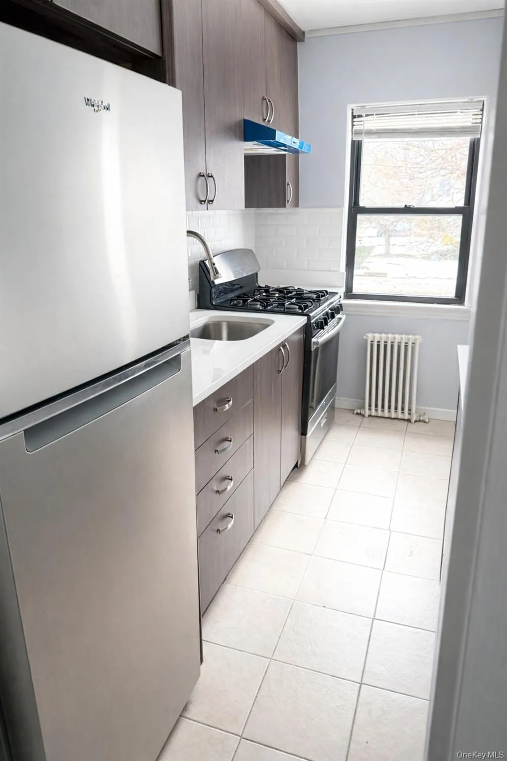 Kitchen with stainless steel appliances, radiator, light tile patterned flooring, modern cabinets, and under cabinet range hood Kitchen with stainless steel appliances, radiator, light tile patterned flooring, modern cabinets, and under cabinet range hood
