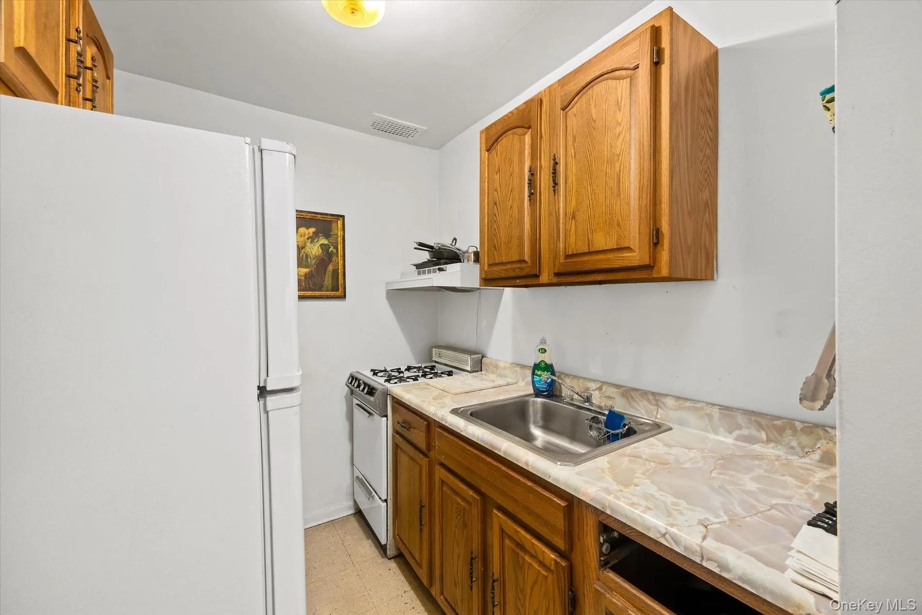 Kitchen featuring wood-finish cabinetry, solid surface countertop, stainless steel sink, and white appliances Kitchen featuring wood-finish cabinetry, solid surface countertop, stainless steel sink, and white appliances