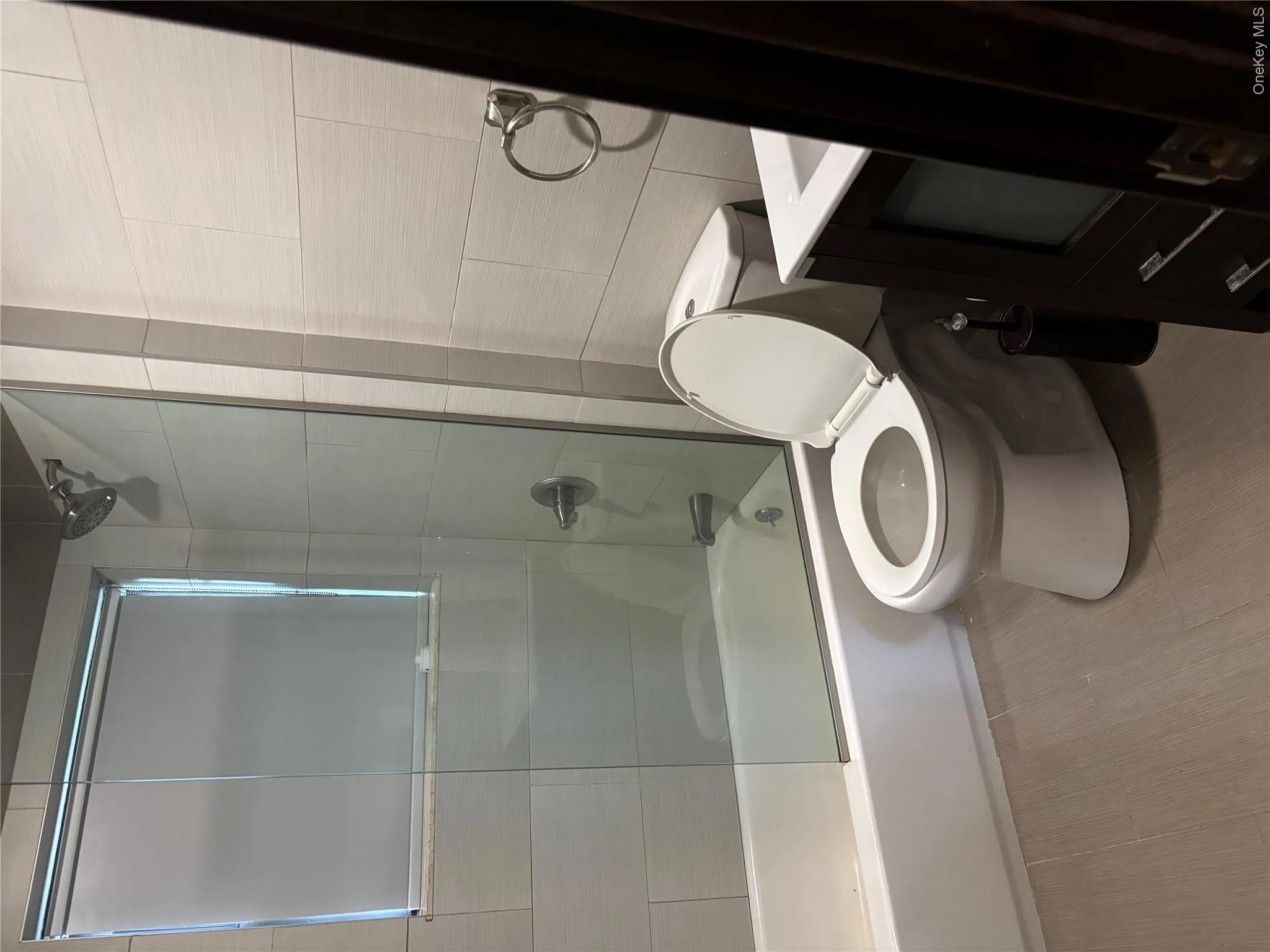 Full bathroom featuring a tub with glass shower enclosure, a toilet, a single vanity with storage, and neutral-toned wall and floor tiling Full bathroom featuring a tub with glass shower enclosure, a toilet, a single vanity with storage, and neutral-toned wall and floor tiling