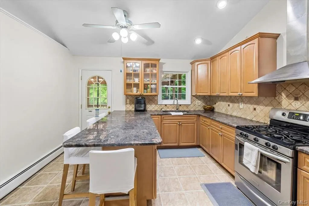 Kitchen featuring stainless steel gas range, a baseboard heating unit, wall chimney range hood, a kitchen breakfast bar, and tasteful backsplash Kitchen featuring stainless steel gas range, a baseboard heating unit, wall chimney range hood, a kitchen breakfast bar, and tasteful backsplash